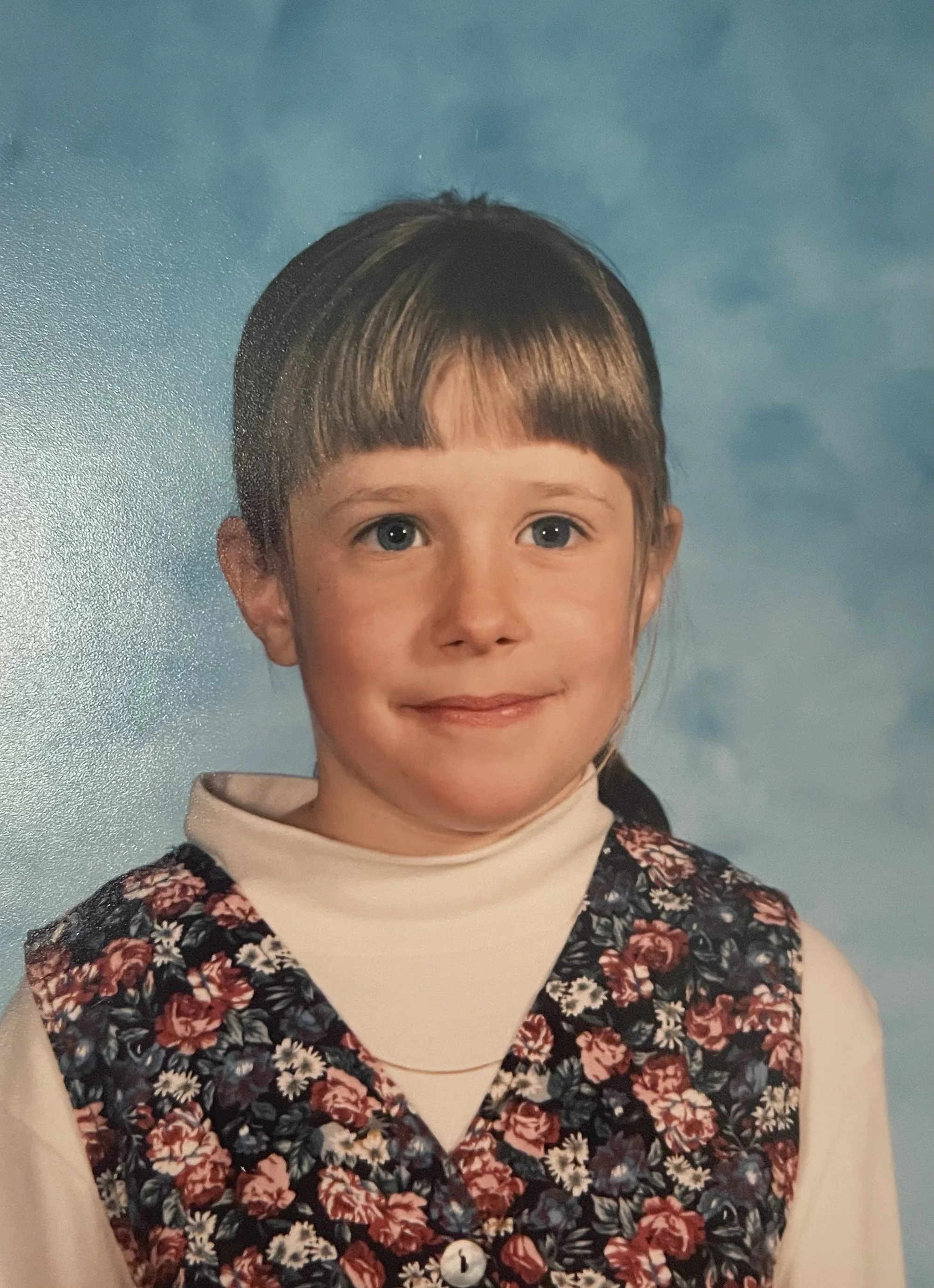A 90s-esk school photo of a young girl, probably 8 years old wearing a floral vest and cream turtleneck. She has bangs and is before a sky blue backdrop.