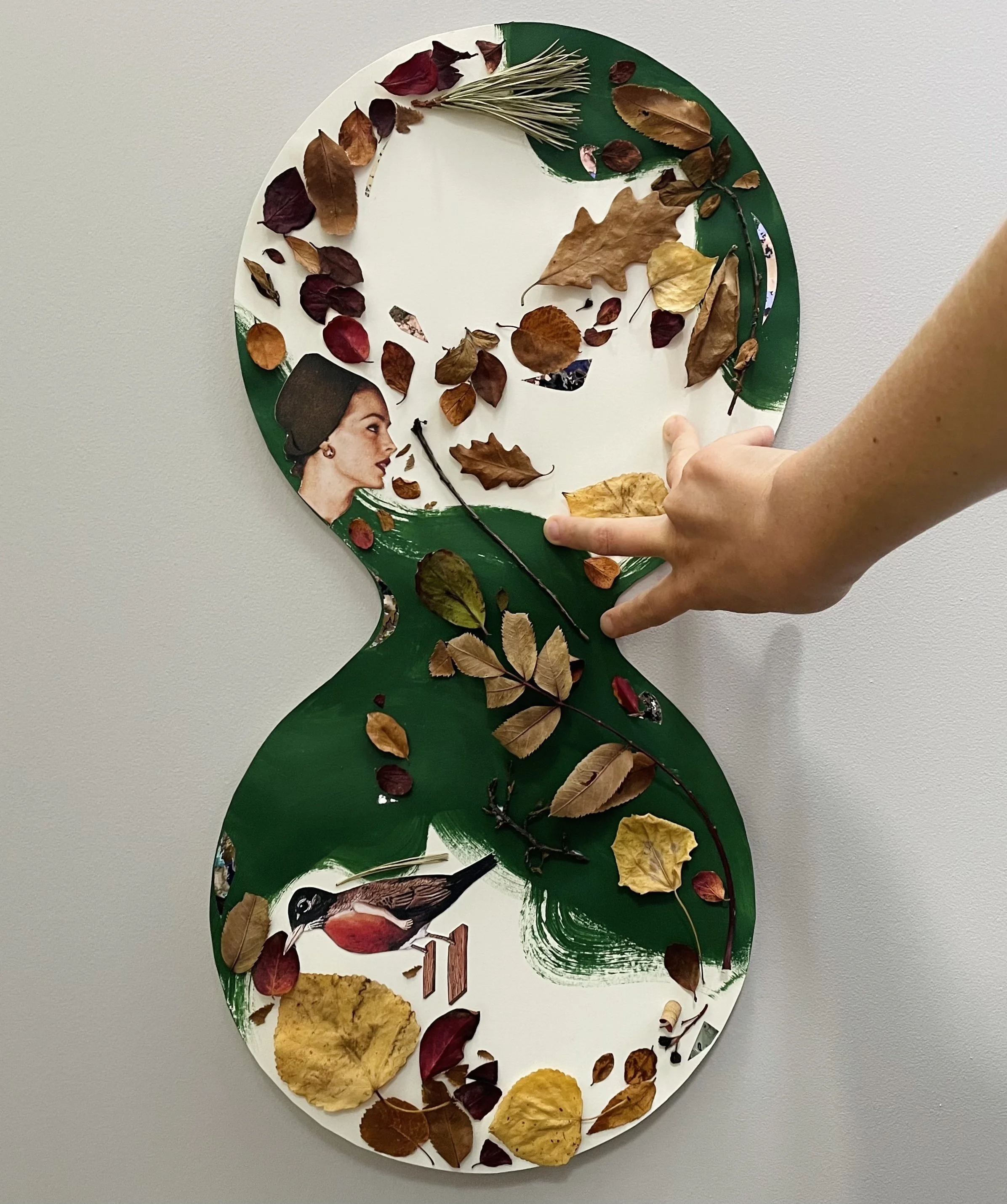 A woman's hand holds a piece of art shaped like a figure eight. On it are dried autumn leaves glued to the green and white painting paper. It also features a 1950s woman and a red-chested bird with wooden planks for feet. Surreal meets nature.