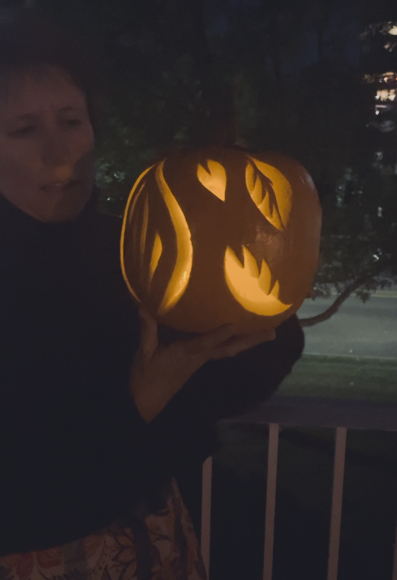 A photo of a carved pumpkin in the dark, held by a woman. It is illuminated. This side of the pumpkin shows different artistic expressions of leaf cutouts. The pumpkin is sprouting nature. 