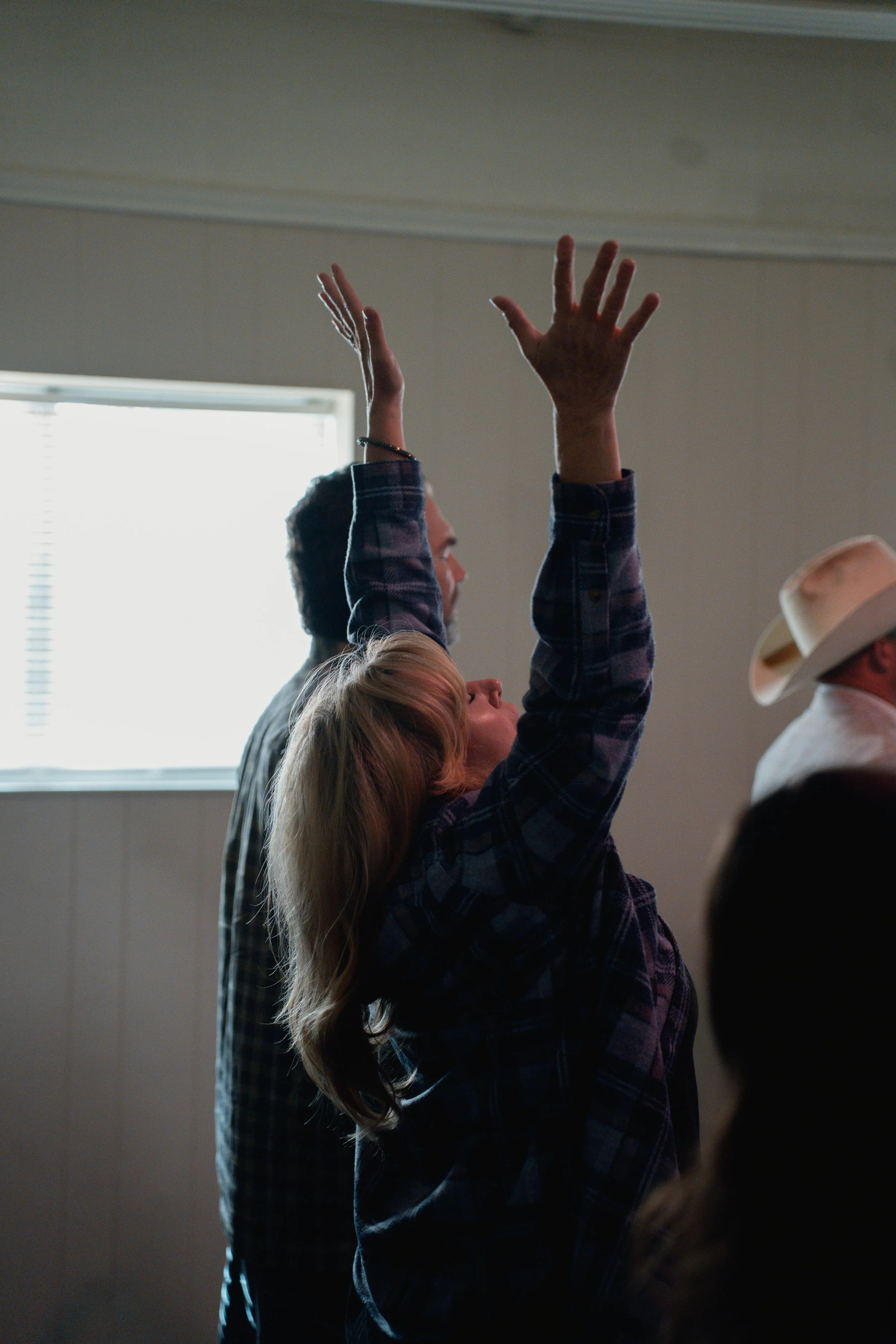 A woman with blonde hair raising her arms in a room with natural light from a window, with two men nearby, one wearing a hat.