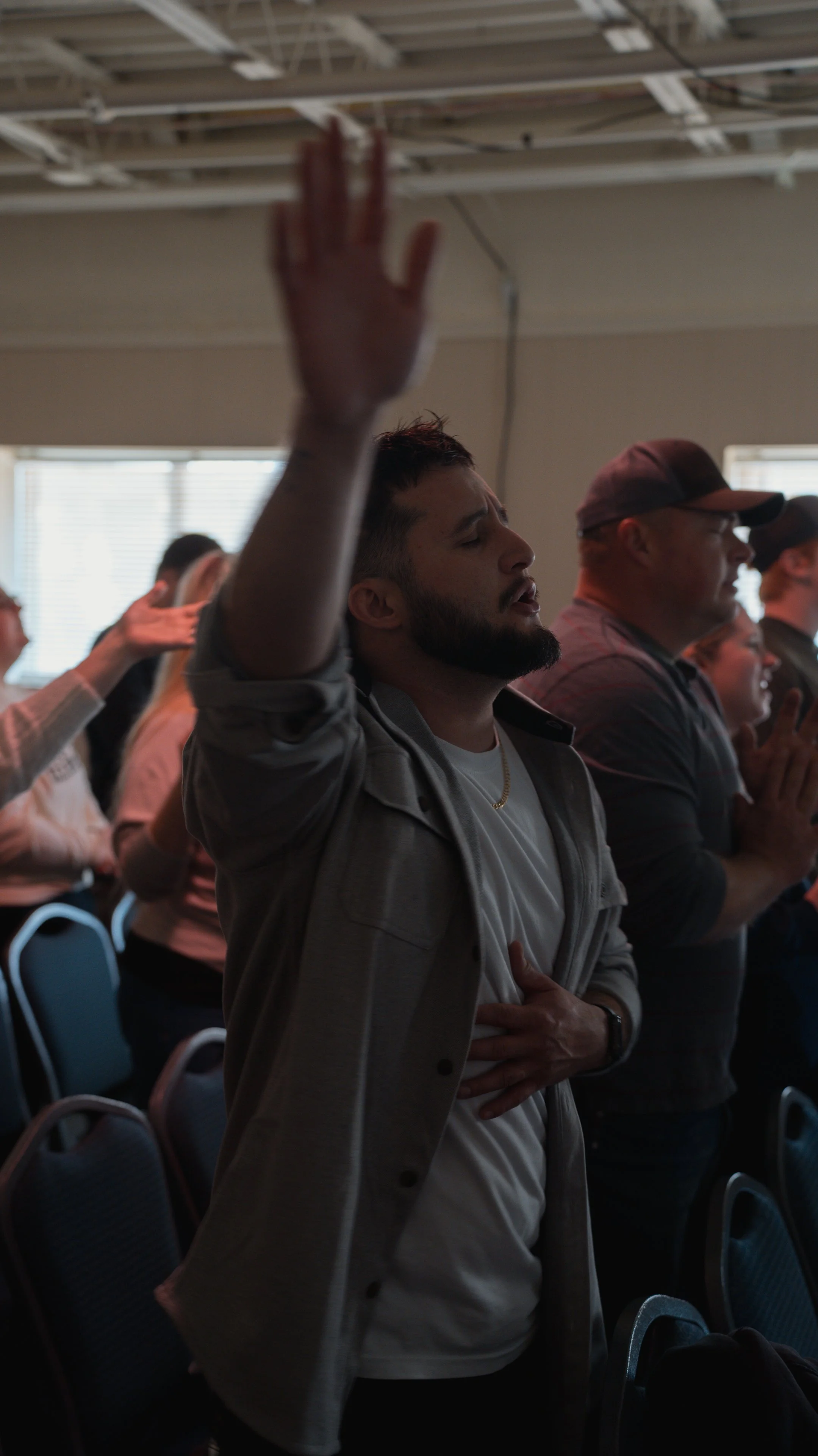 A group of people standing with their eyes closed and hands raised, participating in a spiritual or religious activity in a room with windows.