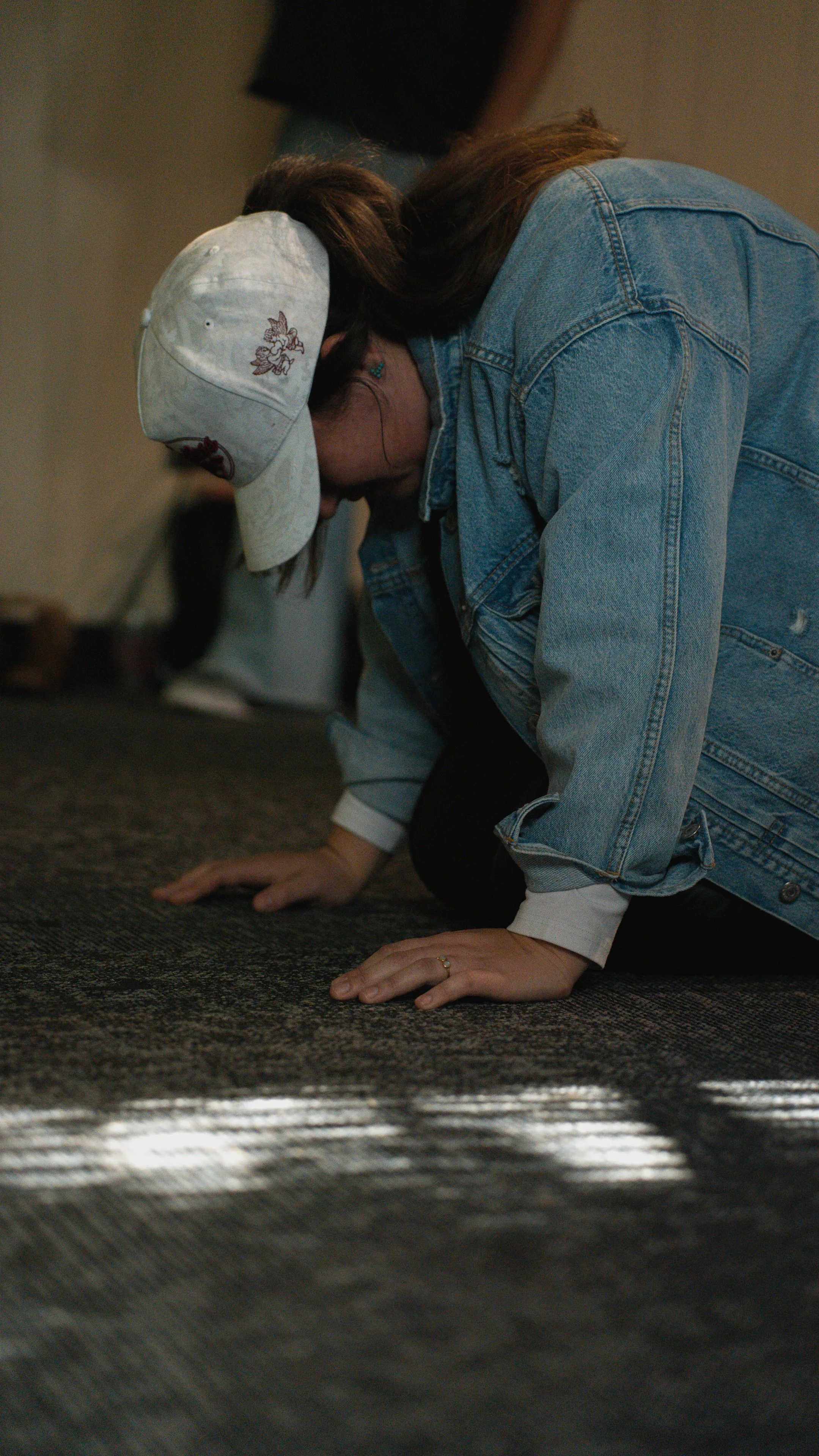 A woman kneeling on the floor with her head bowed down, wearing a light-colored baseball cap and a denim jacket, possibly praying or in a moment of reflection.