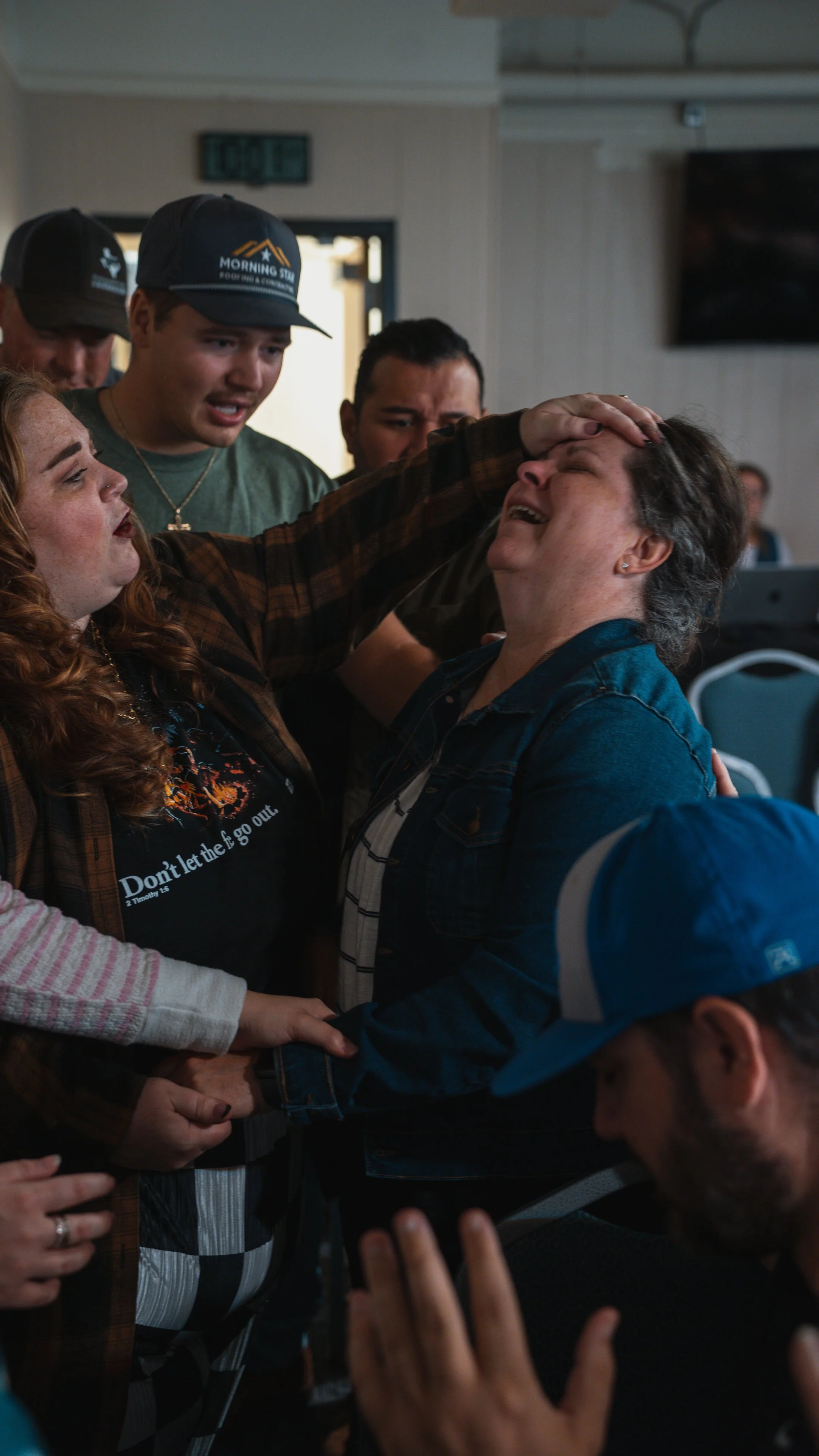 A woman is crying and being comforted by a group of people at an indoor gathering, some holding her hands and touching her head in an emotional moment.