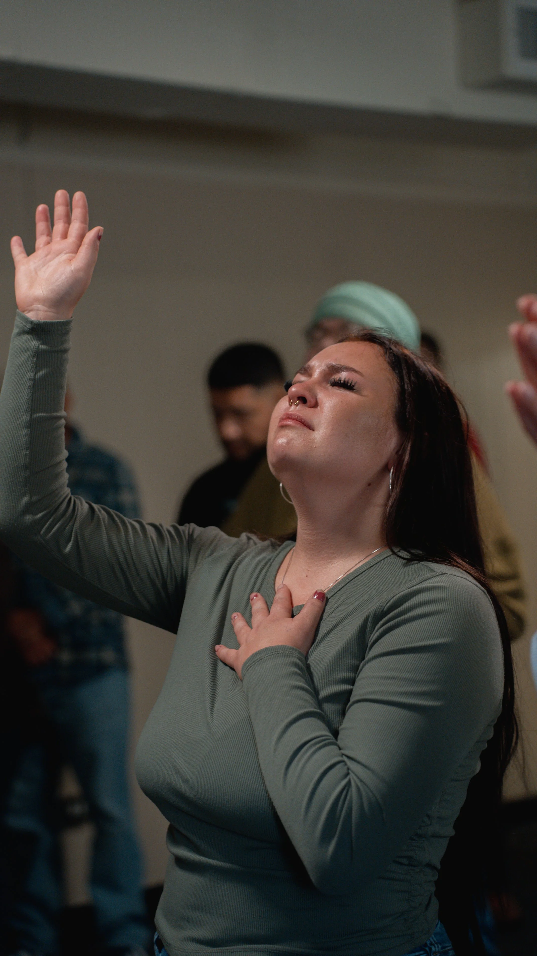A woman with her eyes closed, raising one hand and placing her other hand on her chest, appearing to be praying or worshiping in a room with other people in the background.