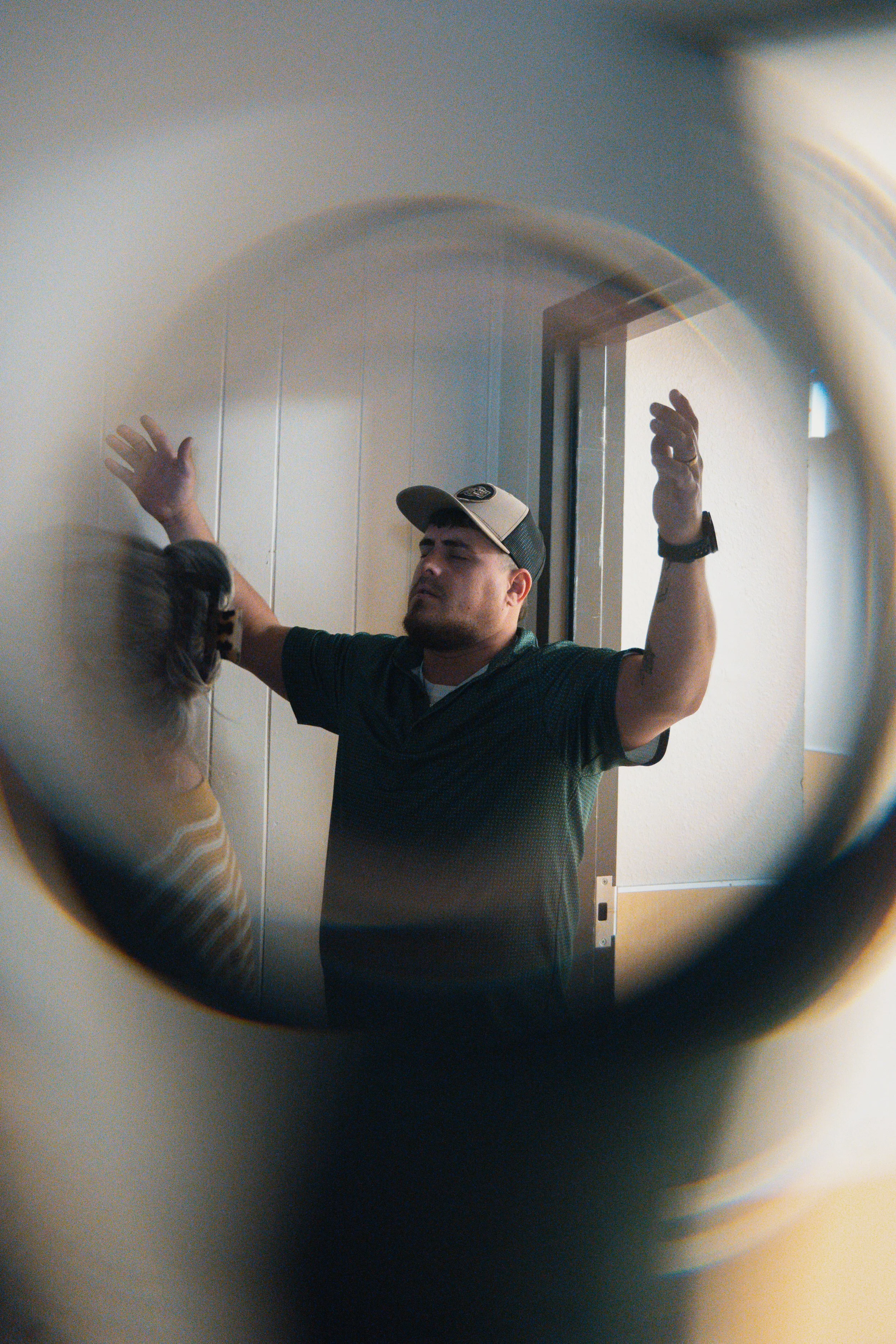 Person with closed eyes and raised hands in a room, viewed through a circular opening.