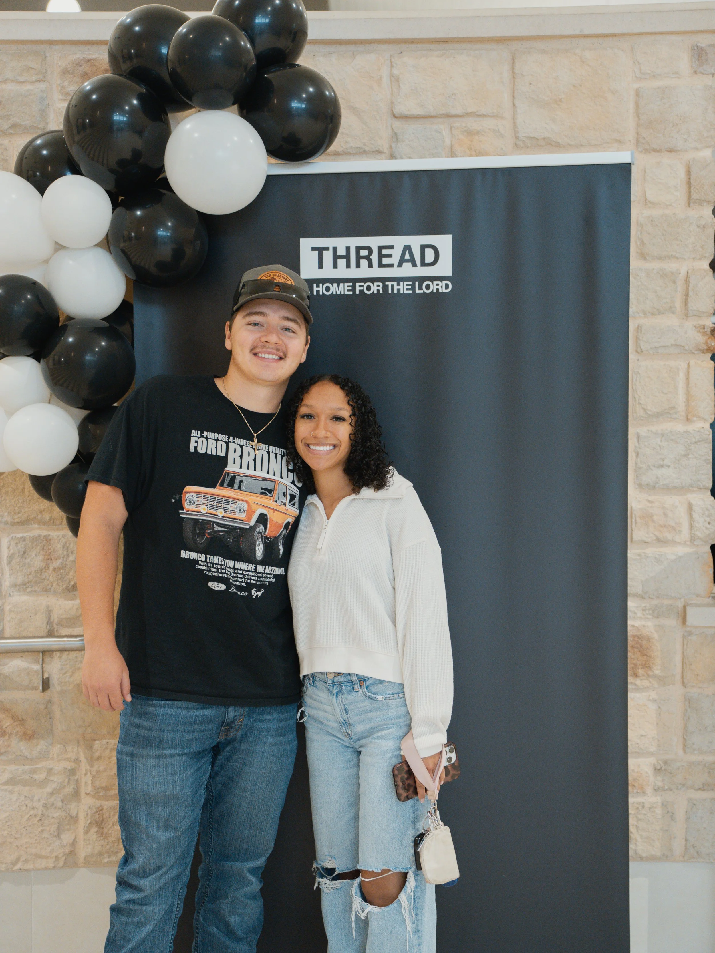 A young man and woman smiling and posing together in front of a dark blue backdrop with the logo 'THREAD' and the slogan 'HOME FOR THE LORD', with black and white balloons arranged on the left side.