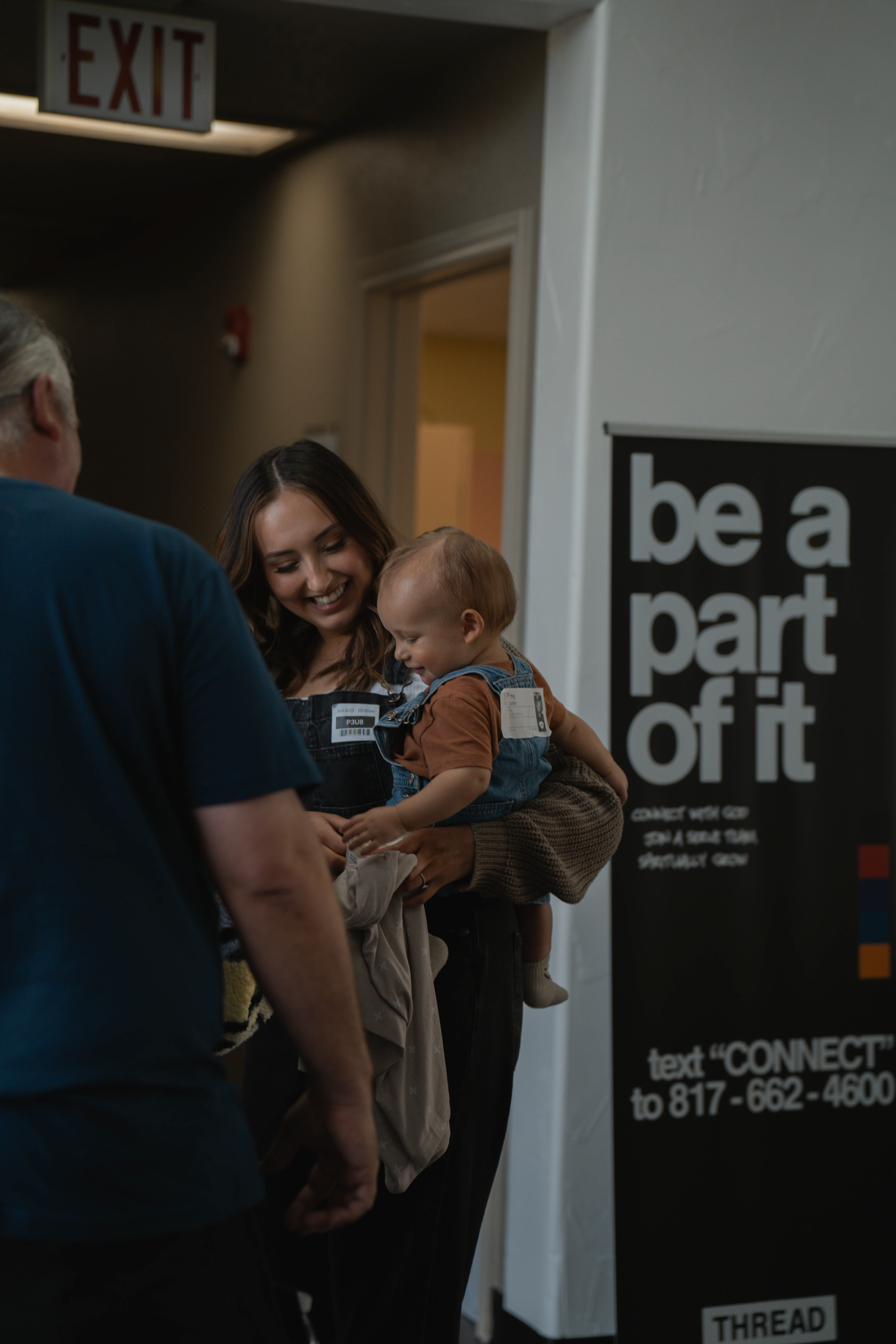 A woman holding a smiling toddler girl in her arms while talking to a man, near a sign that reads 'be a part of it'.
