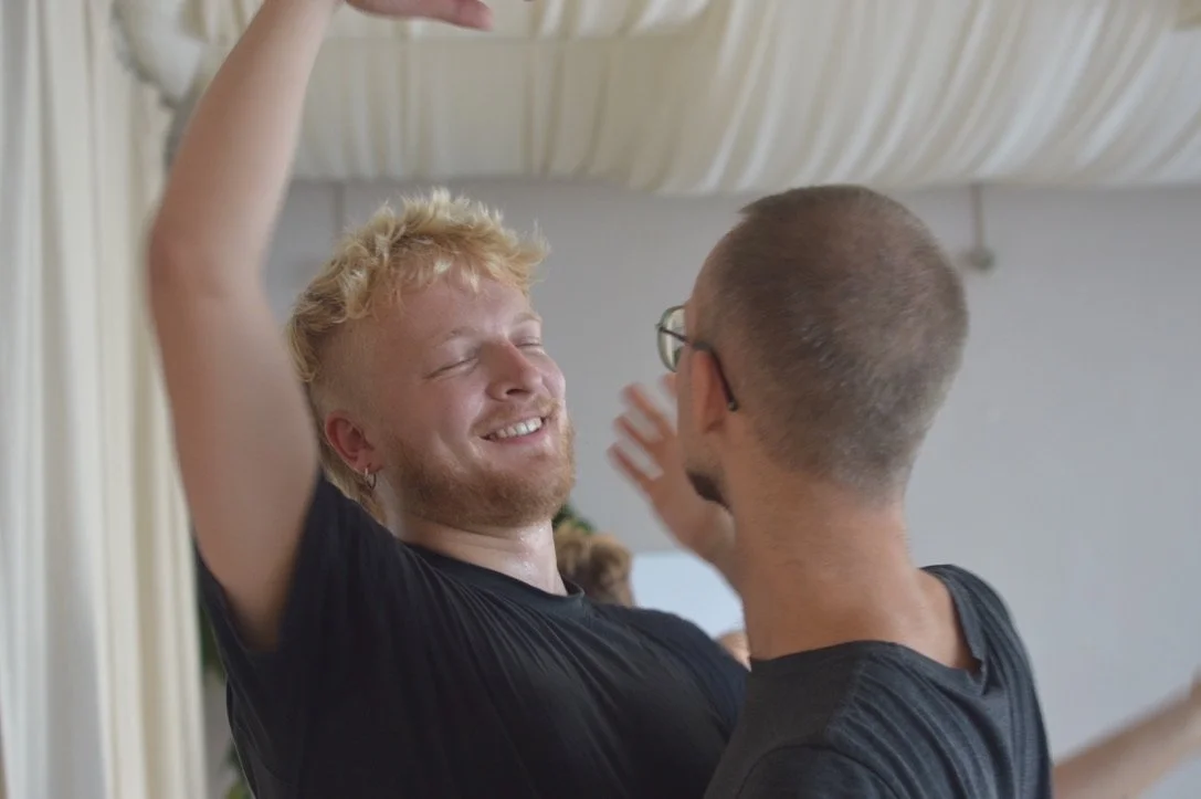 Two men are joyfully dancing together indoors, with one raising his arm and smiling.