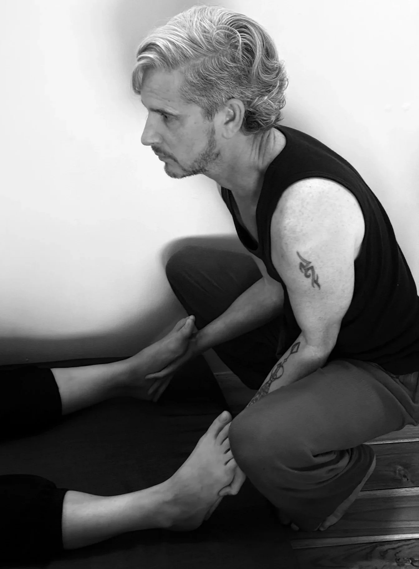 Simon Magnus supporting a participant. He is kneeling on the floor, holding a person's foot and leg, who is lying on a mat, with their toes pointing away from him.