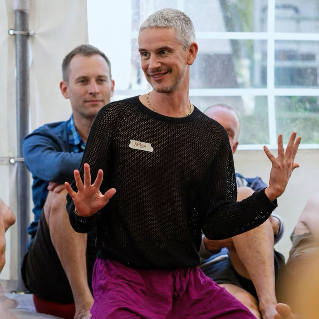 Simon Magnus wearing a black mesh shirt and purple trousers, facilitating a workshop with people sitting behind him in an indoor setting with large windows.