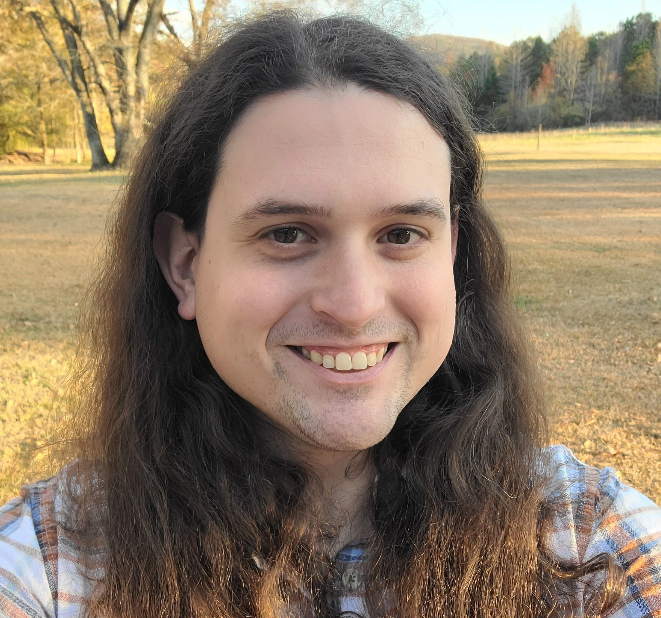 A smiling person with long, wavy brown hair outdoors in a grassy area with trees and hills in the background, during sunset.