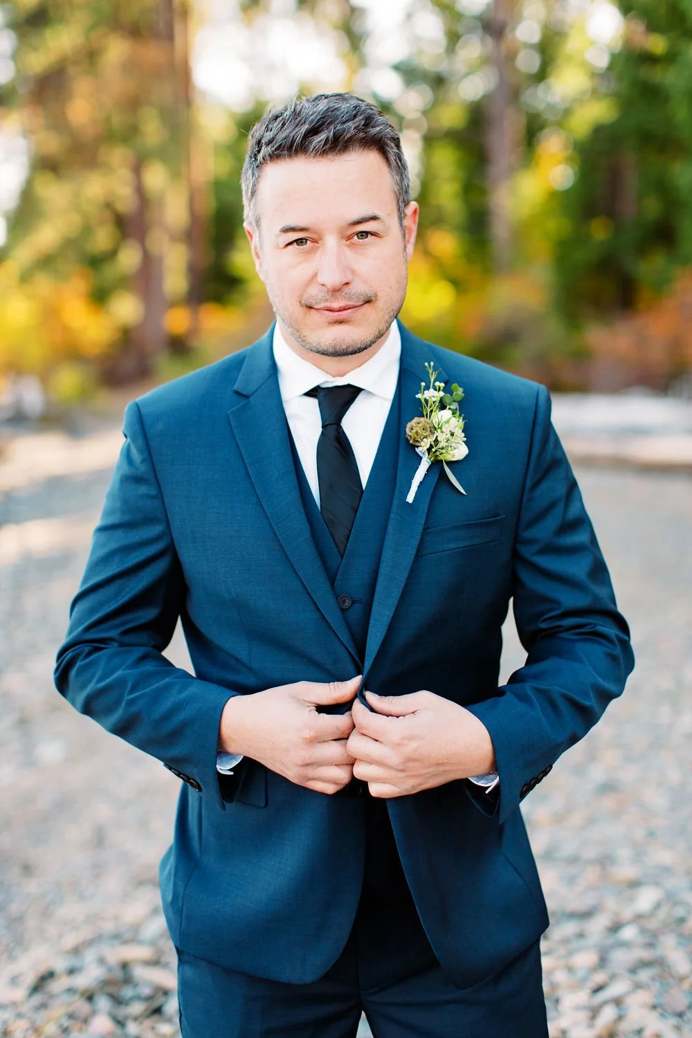A man in a navy blue suit with a boutonniere on the lapel, standing outdoors with a blurred nature background, adjusting his suit jacket.