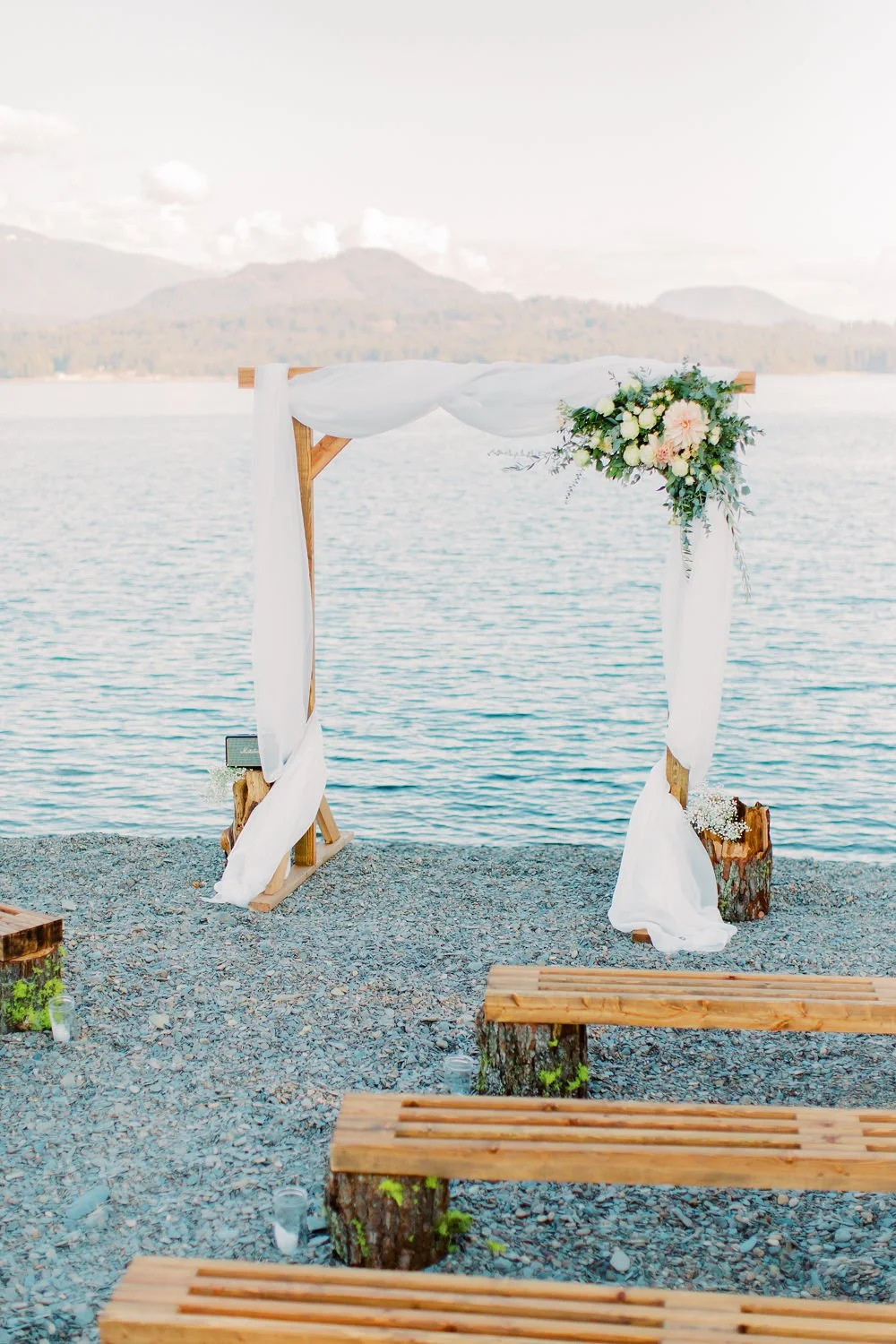 A wedding arch decorated with white fabric and flowers by a lake, with wooden benches on a rocky shore in the foreground and mountains in the background.
