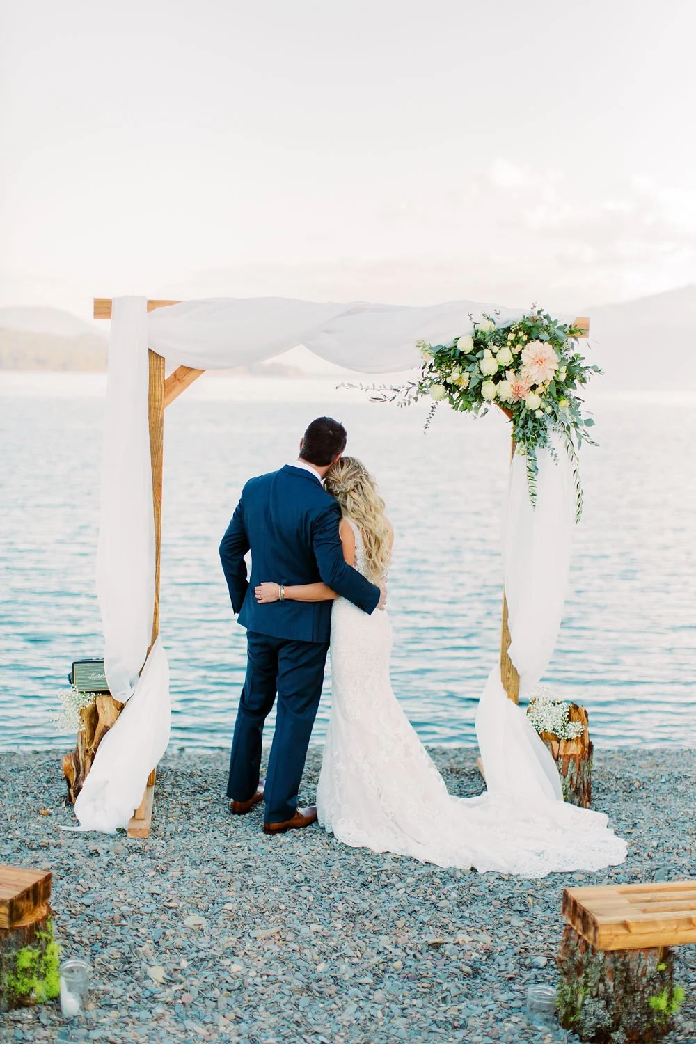 A bride and groom embrace under a wedding arch by a lake, with mountains in the background.