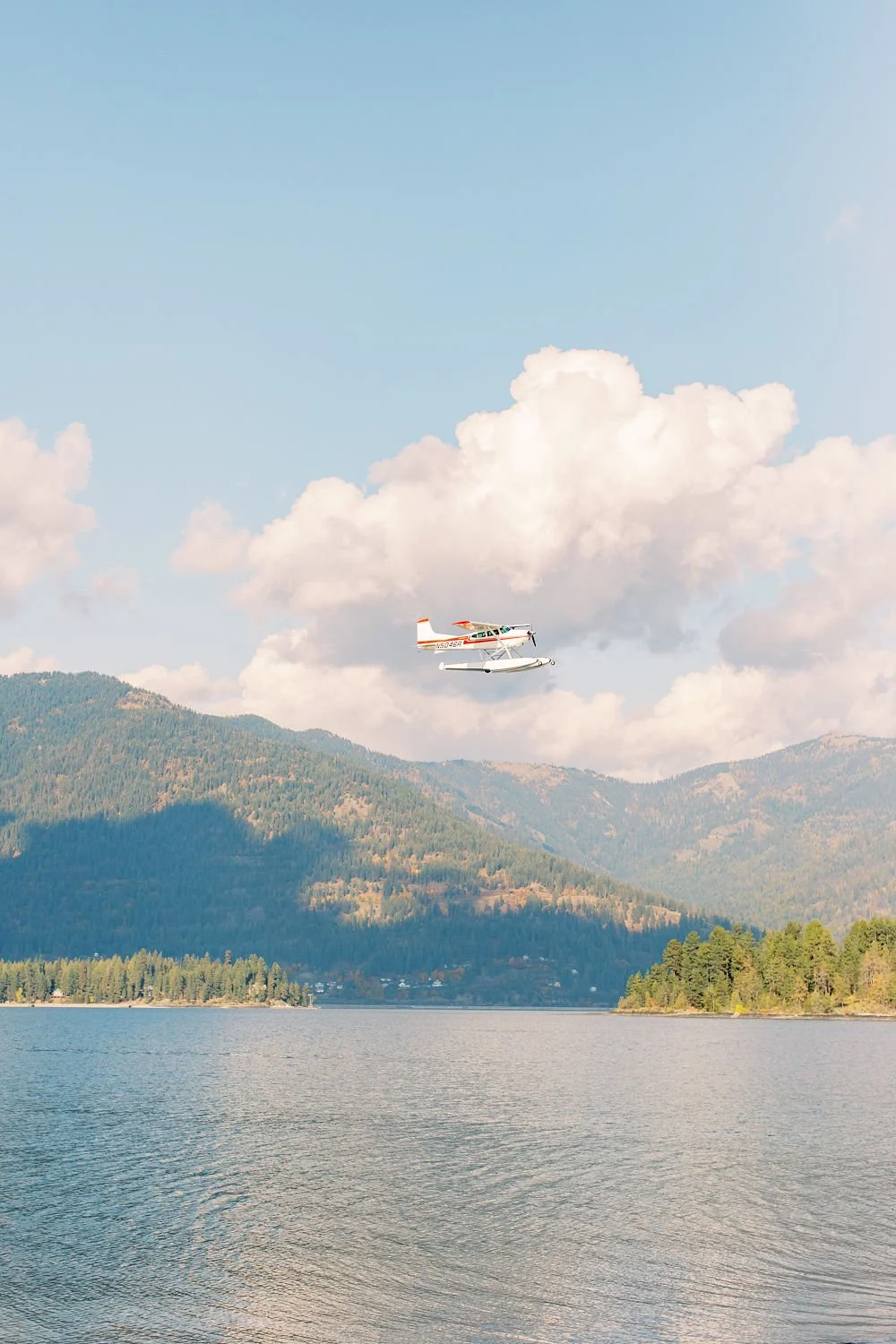 Small airplane flying over a lake with forested mountains in the background under a partly cloudy sky.