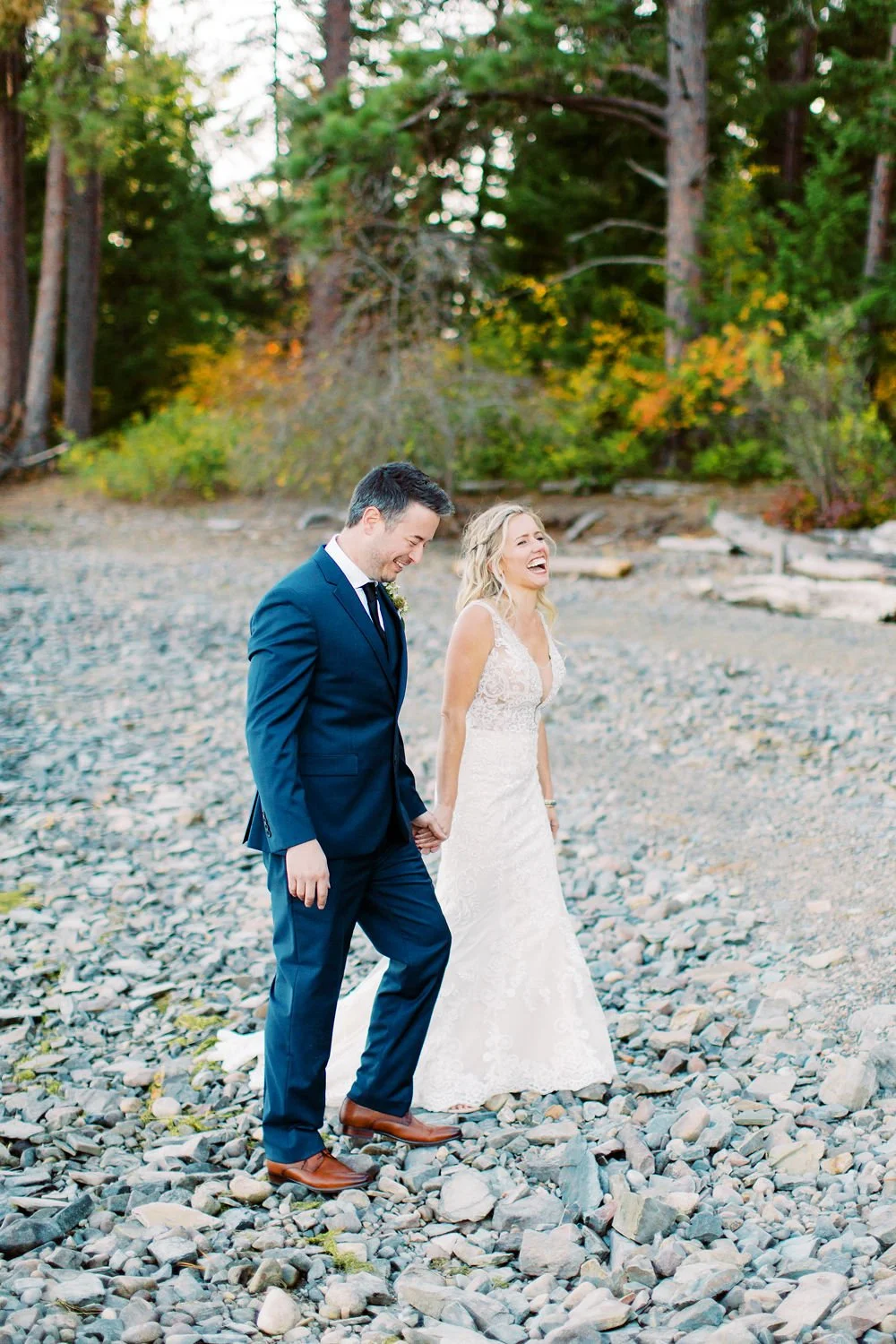 A newlywed couple walking hand in hand on a rocky beach, laughing and enjoying each other's company during sunset with trees in the background.
