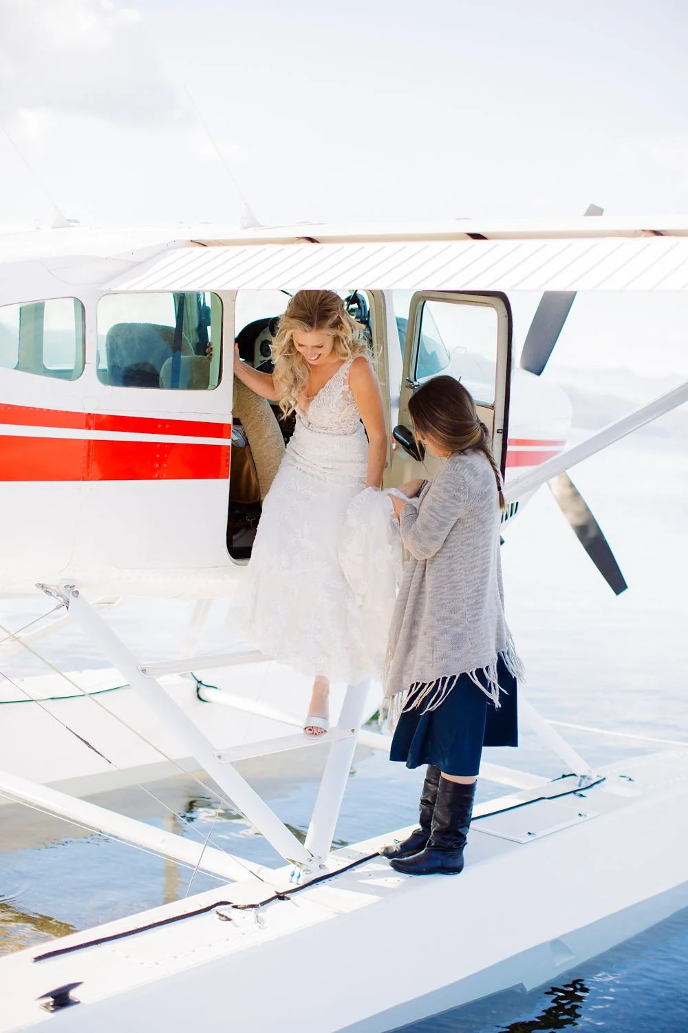 A woman in a white wedding dress getting into a seaplane with assistance from a woman in a gray sweater and black boots.