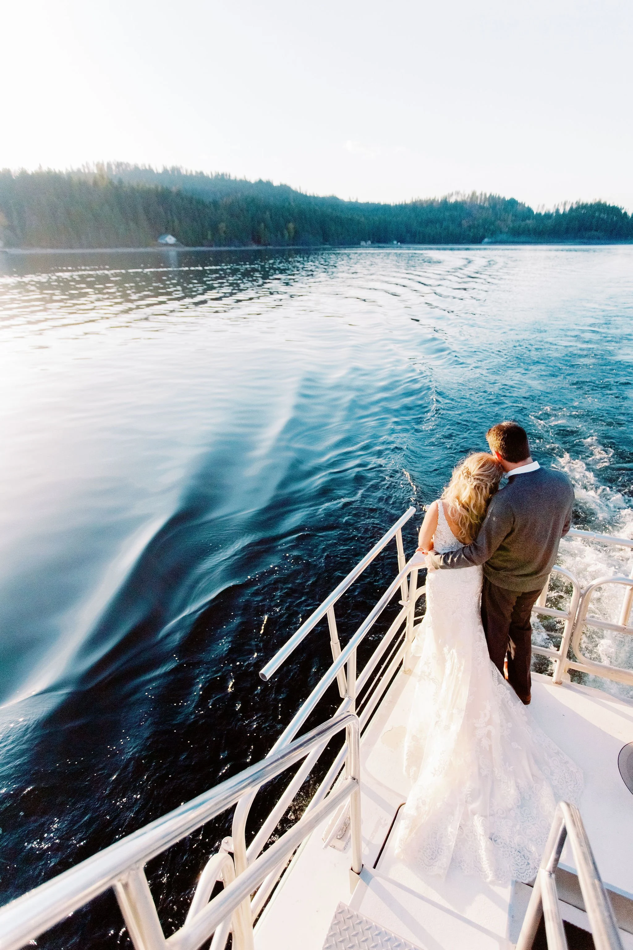 Bride and groom embracing on a boat at sunset on a lake with forested shoreline in the background.