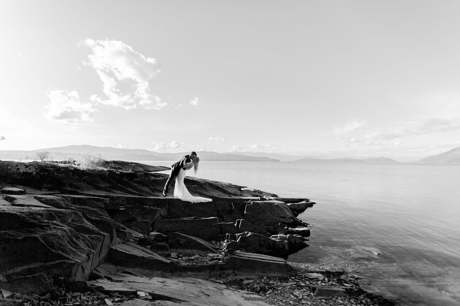 A black and white photograph of a couple on a rocky shoreline, with the man dipping the woman in a dance pose, near the water with mountains and sky in the background.