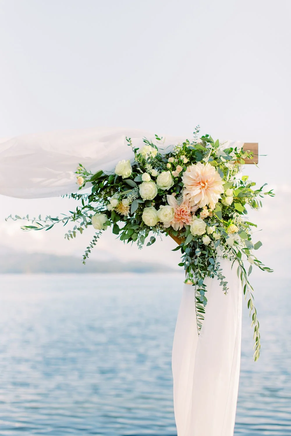 A floral arrangement with white and blush pink flowers, greenery, and flowing white fabric, set against a body of water and sky background.