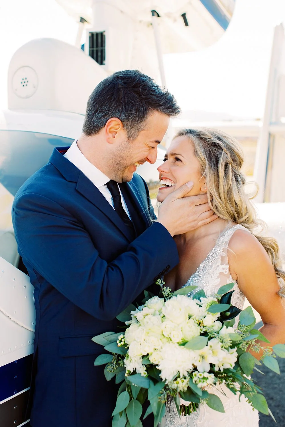 A happy couple in wedding attire sharing a joyful moment outdoors, with the man holding a bouquet of white flowers and greenery.