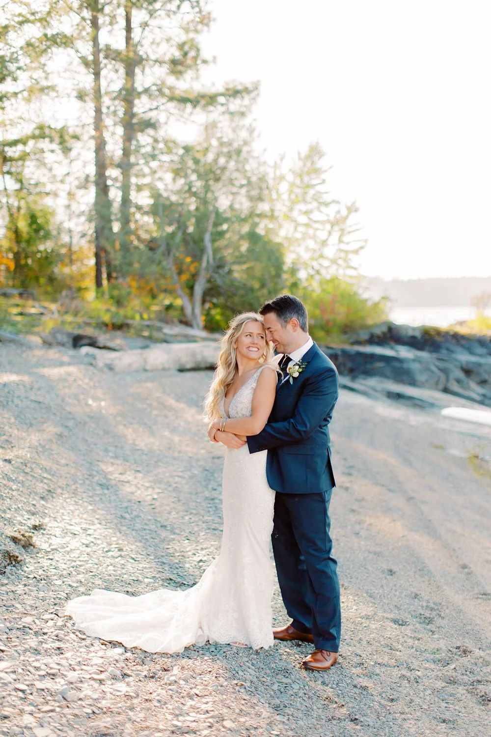 A bride and groom standing close on a rocky beach, smiling and sharing an intimate moment during sunset with trees in the background.