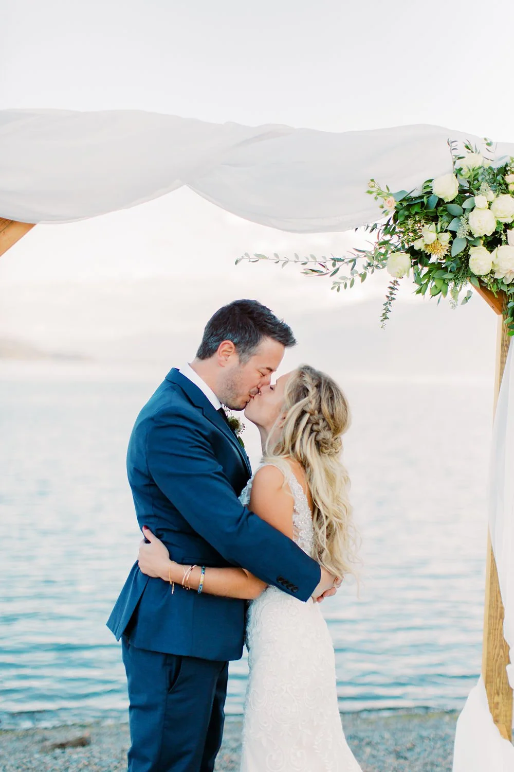 A bride and groom kissing during their wedding ceremony on a beach, decorated with white drapery and a floral arrangement.