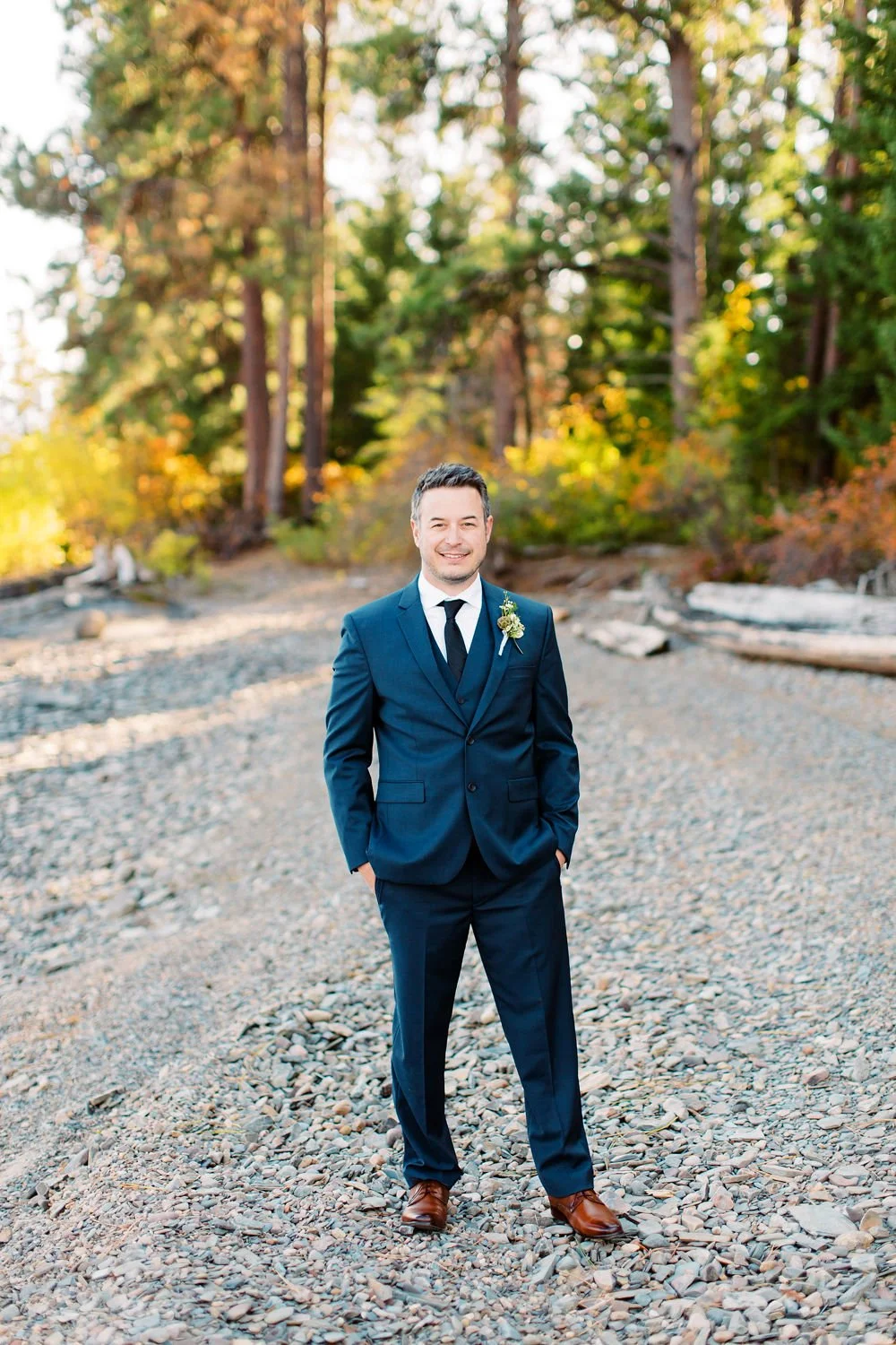 A happy man in a navy blue suit, white shirt, and black tie standing on a rocky outdoor path with trees in the background during autumn.