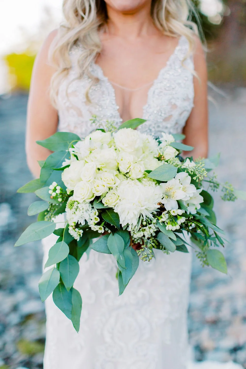 A bride in a lace wedding dress holding a bouquet of white roses and greenery.