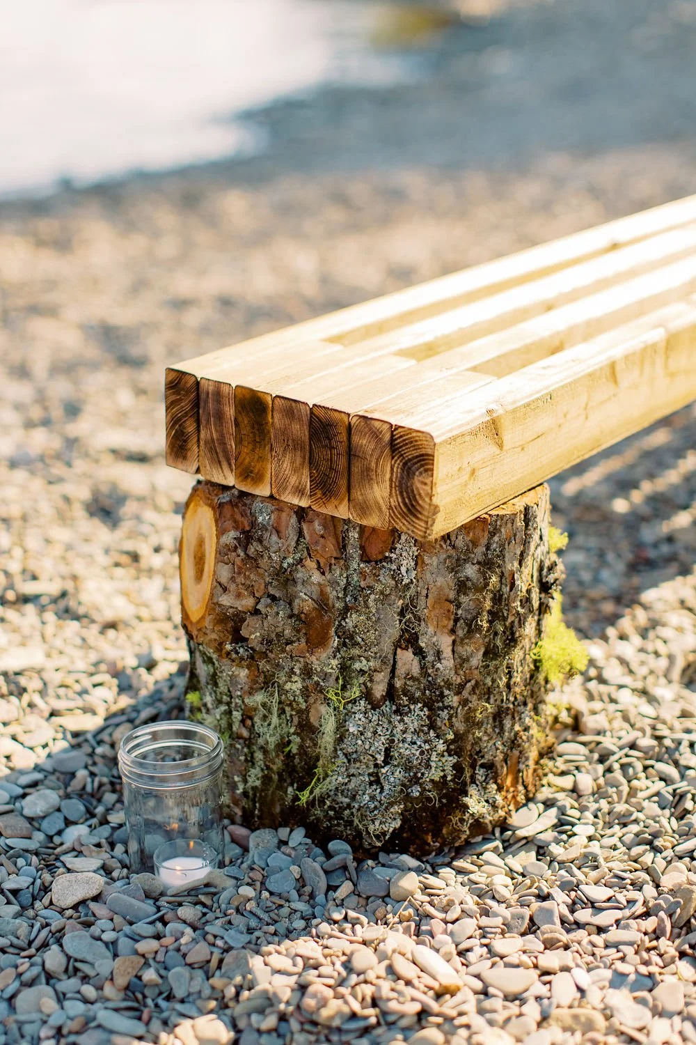 Wooden bench resting on a tree stump on a pebbled beach with a candle in a glass holder nearby, near water.