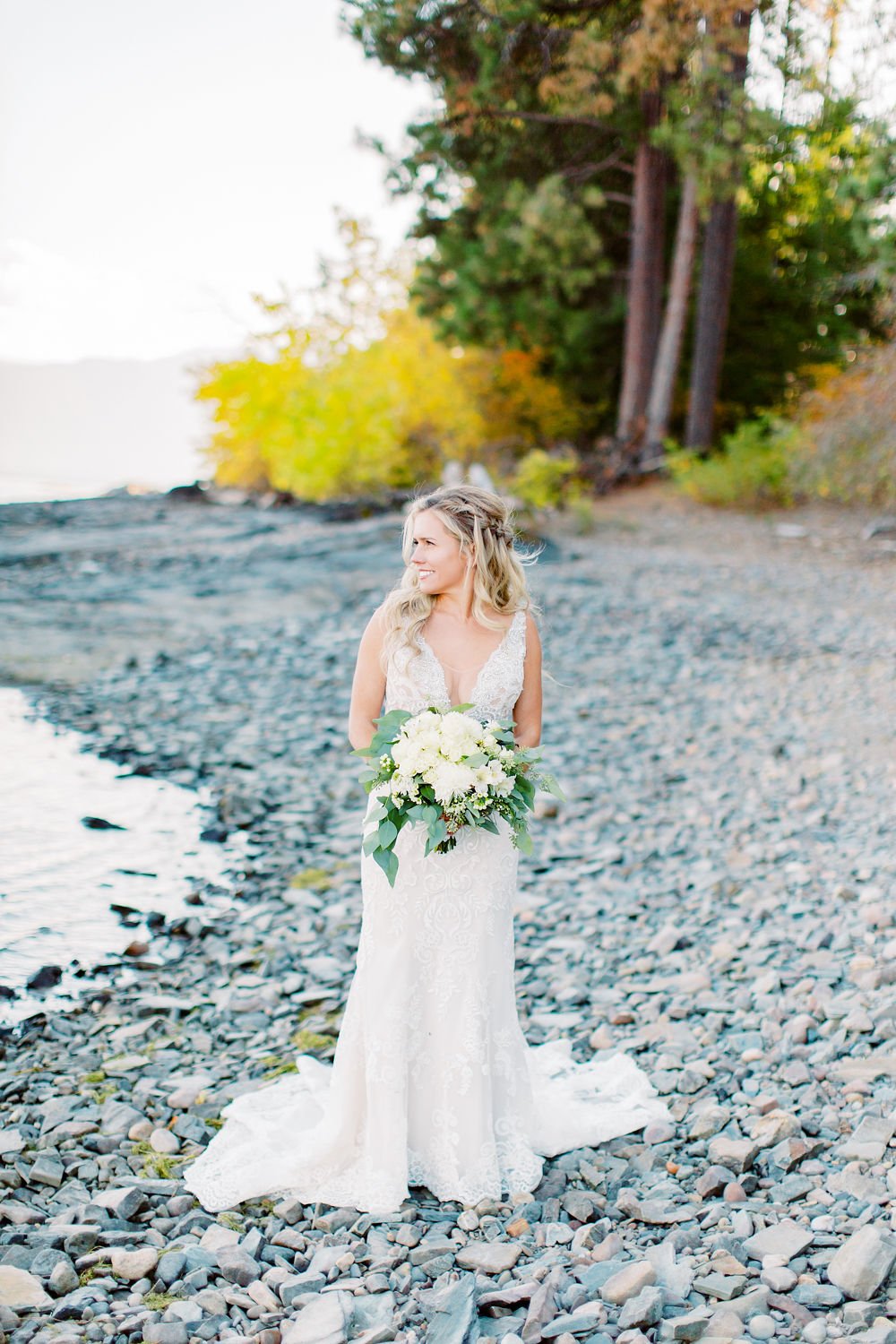 A bride in a white lace wedding gown holding a bouquet of white flowers and greenery on a rocky beach with trees in the background.