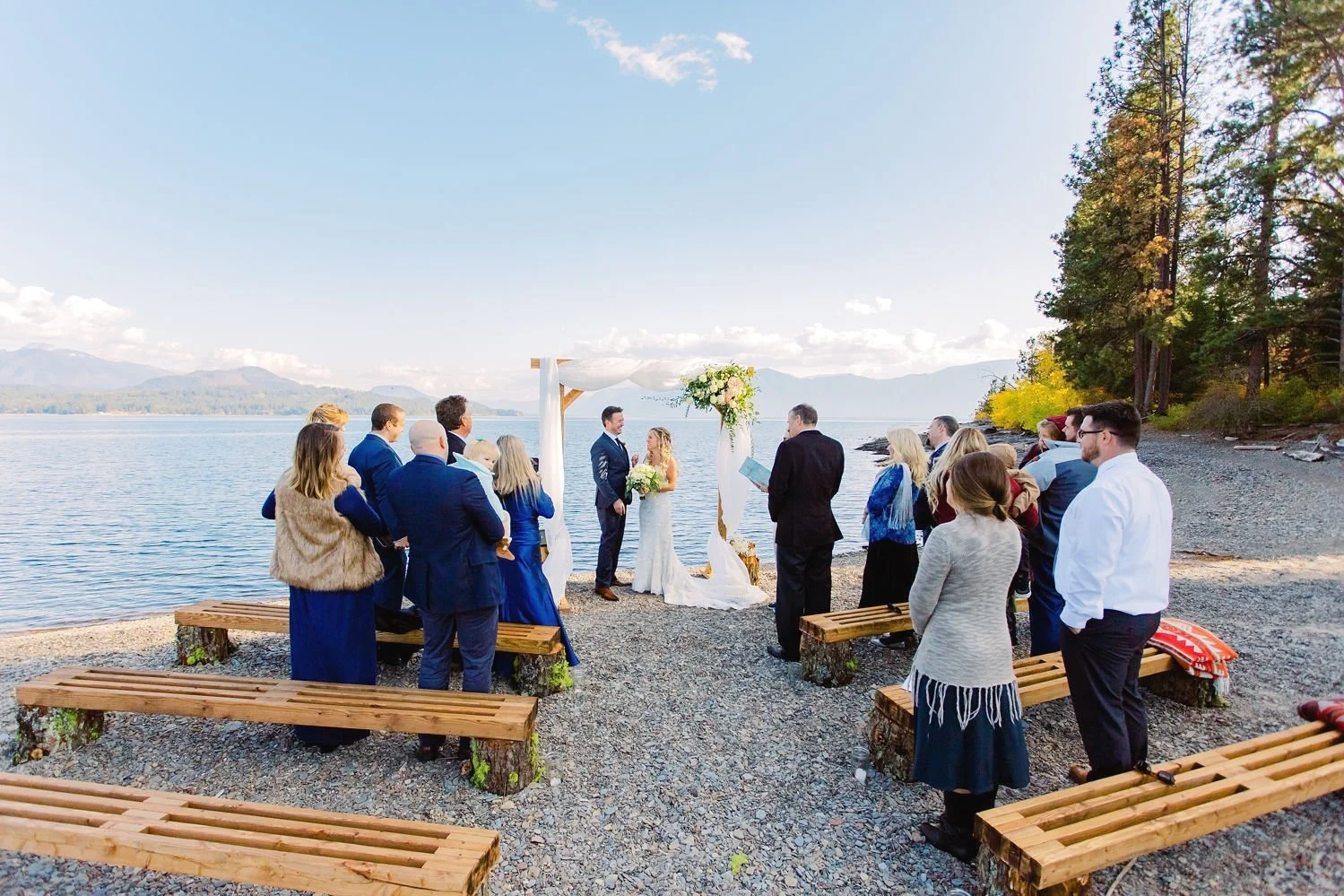 A wedding ceremony taking place outdoors on a rocky beach by the water with trees on the right and distant mountains in the background, featuring a bride and groom standing under a decorated arch with guests around them.