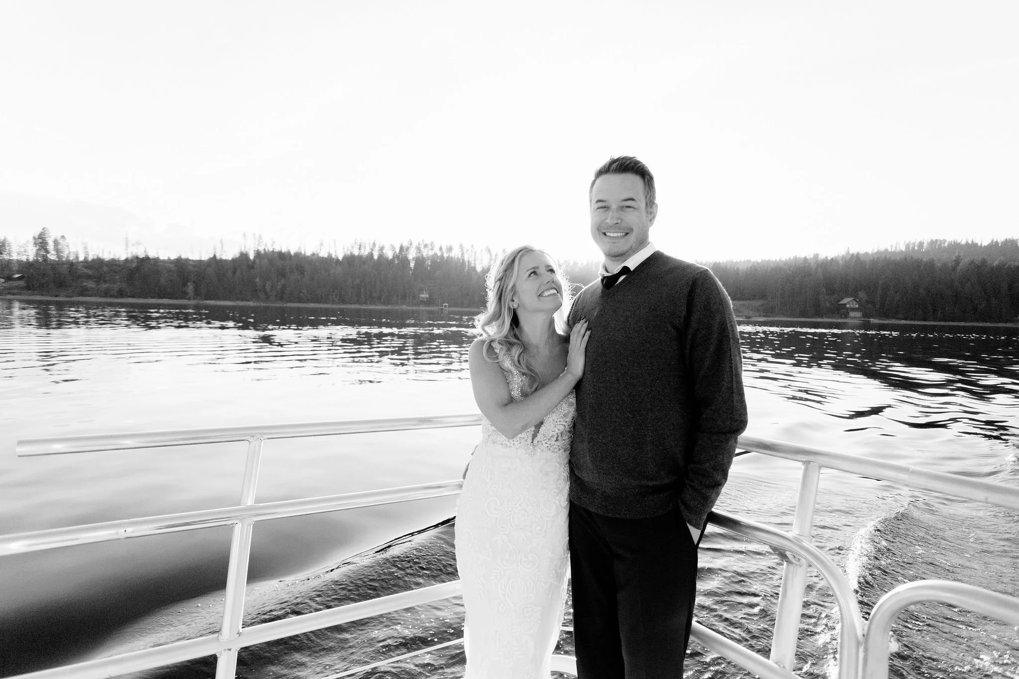 A smiling couple, a woman in a wedding dress and a man in a sweater and shirt, standing on a boat with water and trees in the background.