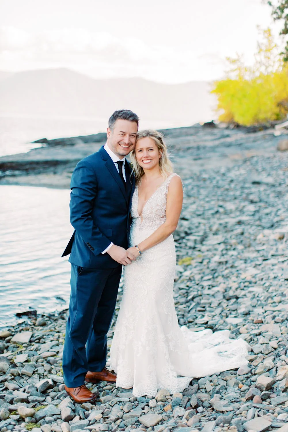 A newlywed couple smiling and holding hands on a rocky beach with water and distant mountains in the background.