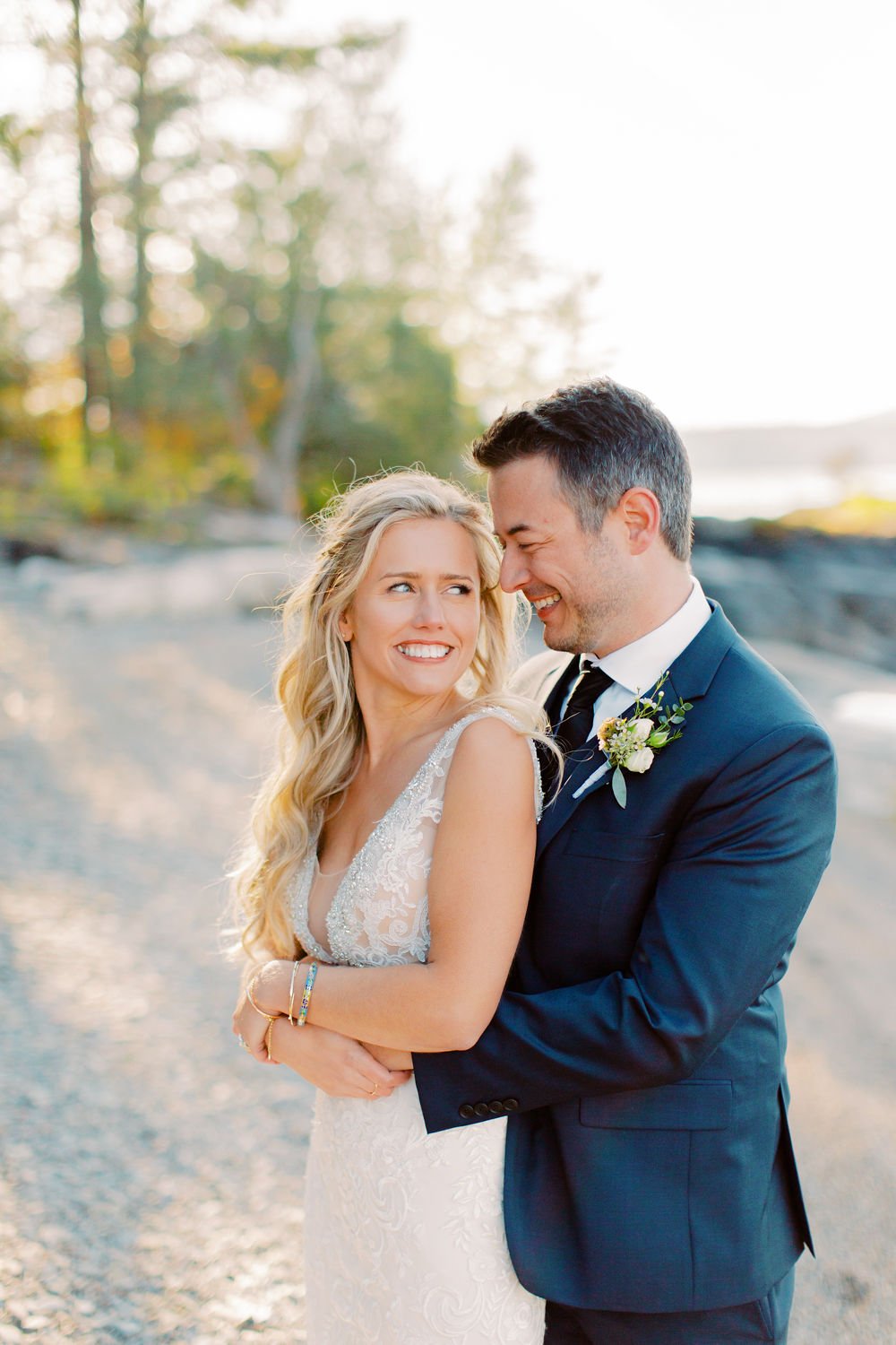 A bride and groom smiling and embracing outdoors on a beach during sunset.