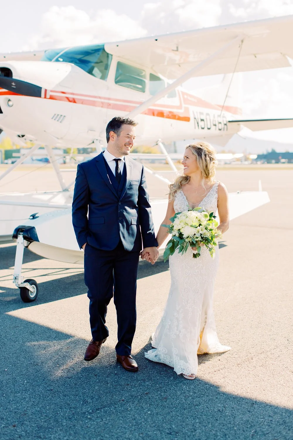 A newlywed couple holding hands and smiling in front of a small airplane on a sunny day.