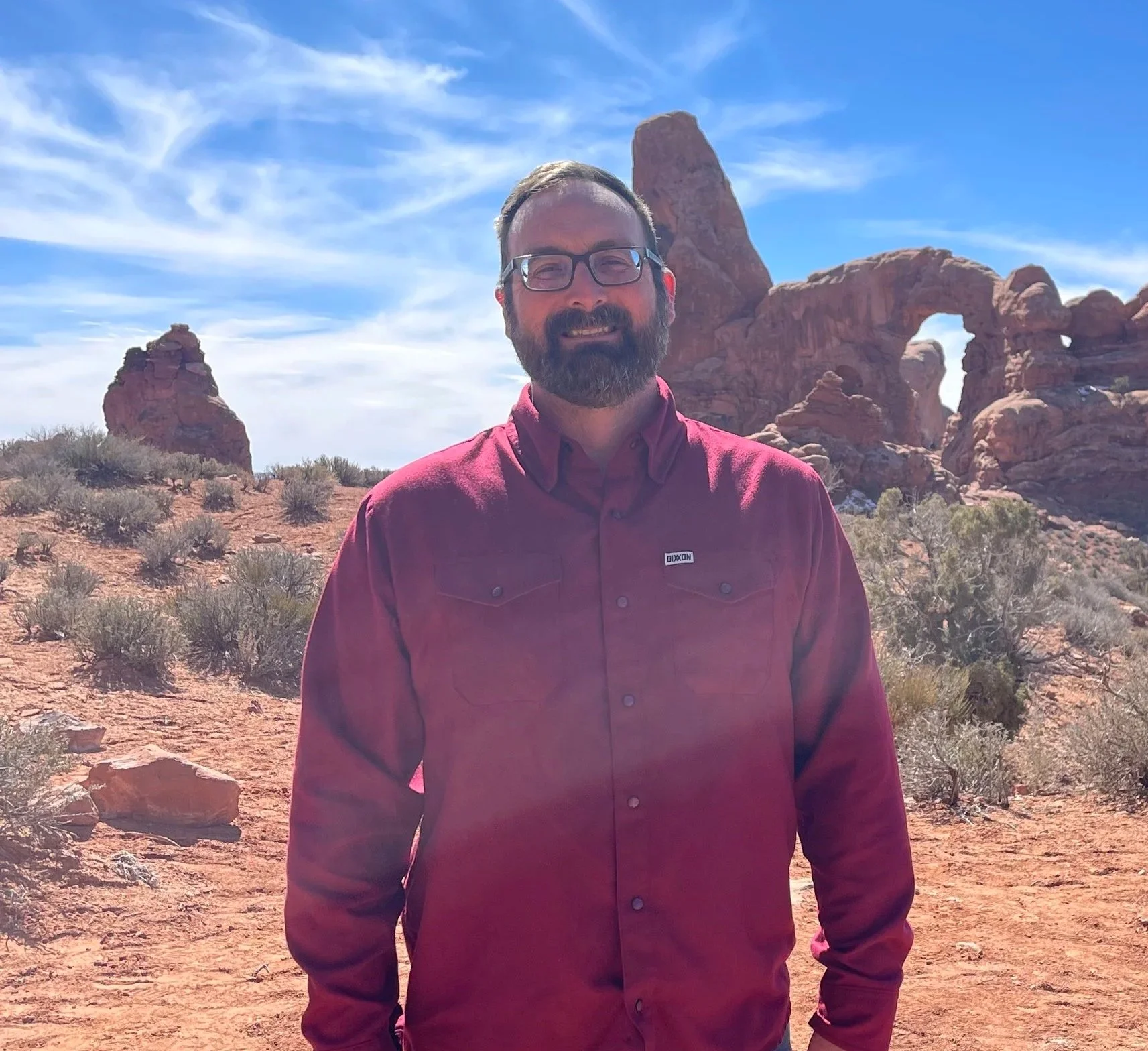 A man with glasses and a beard is standing outdoors in a desert landscape with red rock formations behind him, including a prominent natural arch and large boulder, under a partly cloudy blue sky.