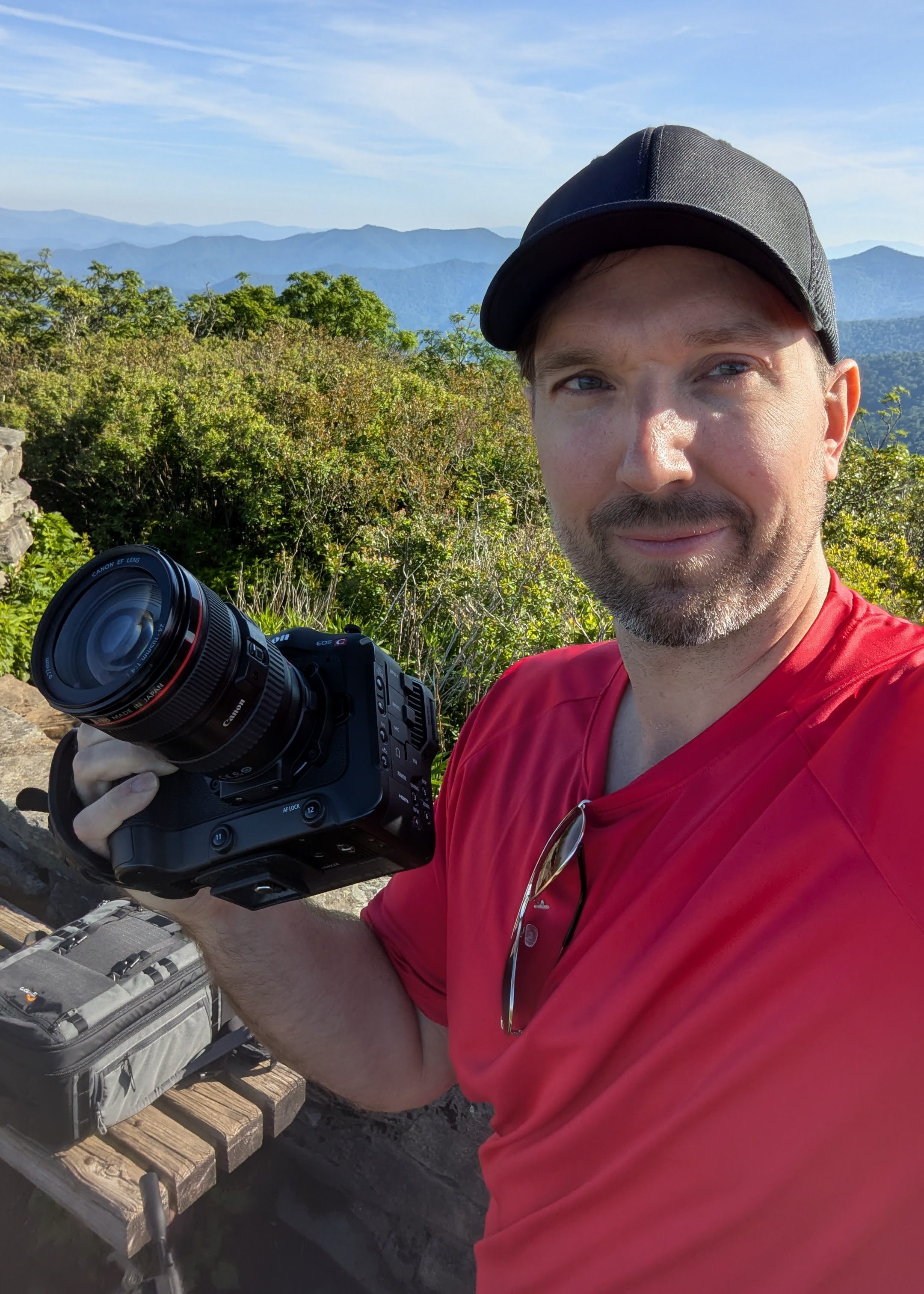 A man wearing a black cap and red shirt taking a selfie with a mountain landscape in the background during daytime. He is holding a camera and standing near a wooden picnic table.