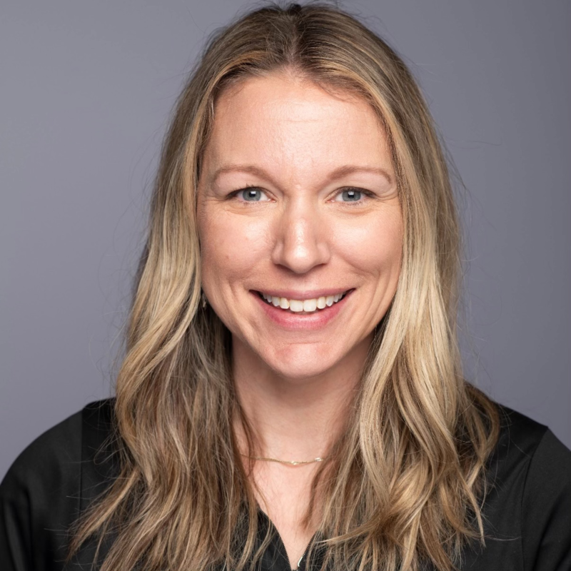 A woman with long, wavy blonde hair, blue eyes, and fair skin smiling at the camera. She is wearing a black top and a delicate necklace against a plain gray background.