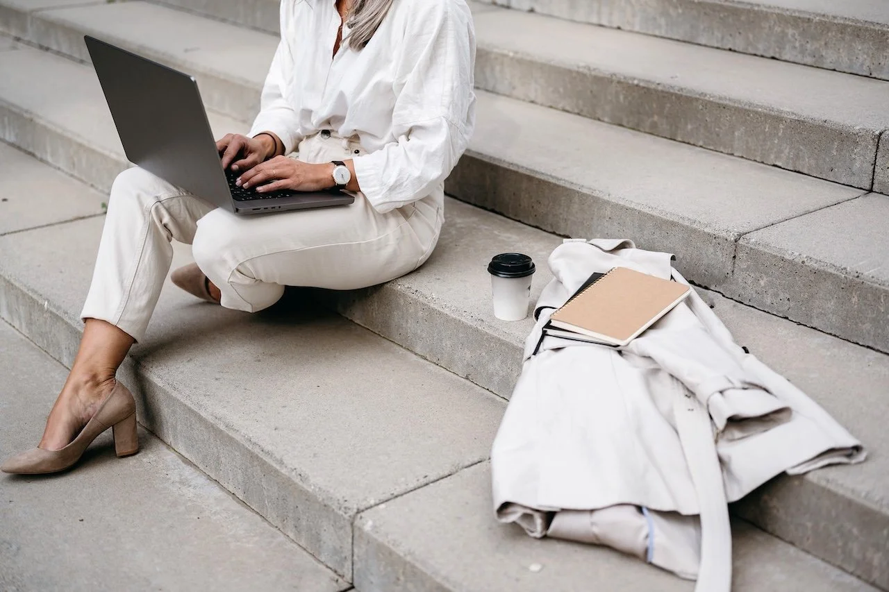 Ein elegante Frau sitzt auf breiten Steinstufen und tippt auf ihren Computer.
