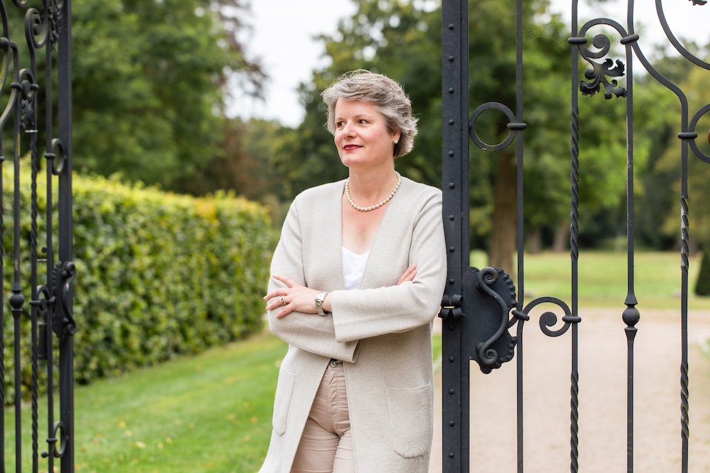 Lydia steht an einem schwarzen Gittertor und blickt nach rechts. Das ist der Eingang zu einem großen, grünen Park.