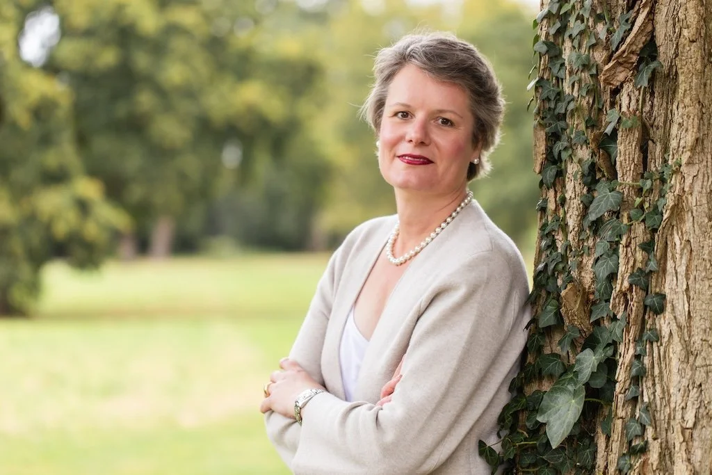 Lydia Böhm lehnt an einem alten, efeubewachsenen Baum, Sinnbild für Herkunft und Generationenmuster in der Finanzarchitektur.