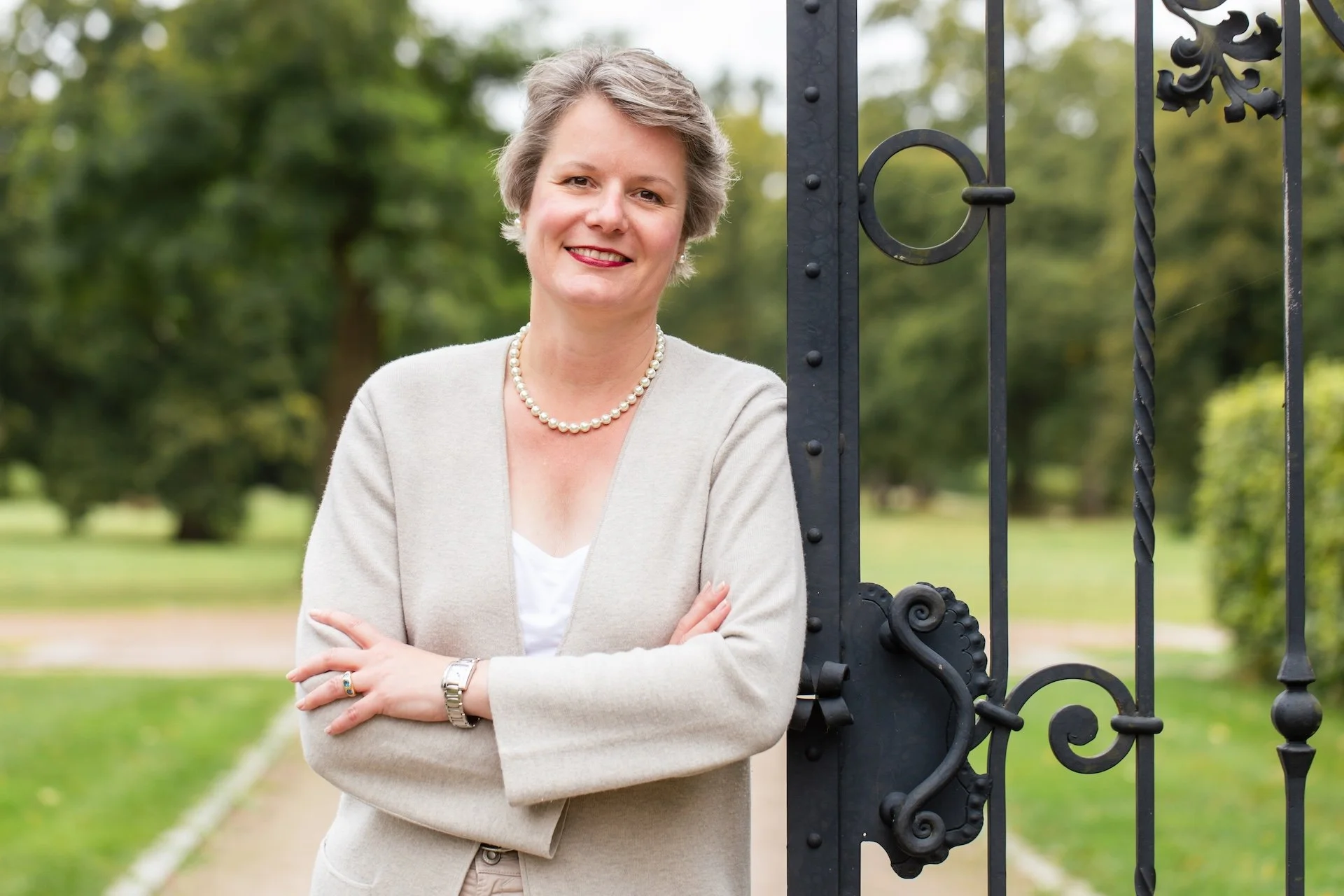 Lydia Böhm, Finanz-Mentorin mit Generationenblick, steht mit verschränkten Armen an einem schmiedeeisernen Parktor und blickt direkt in die Kamera.