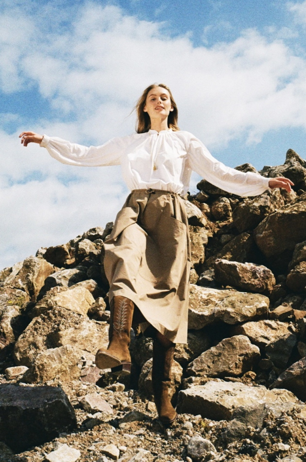 Model wearing Bamford autumn–winter clothing, walking down rocky terrain in a white blouse, neutral skirt and boots under a blue sky.
