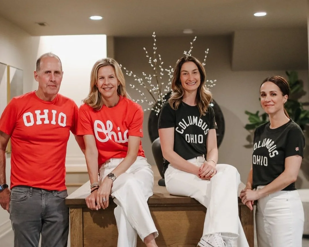 Four people in Ohio-themed shirts smiling indoors, with two sitting on a table and two standing beside it, and a decorative plant with white branches behind them.