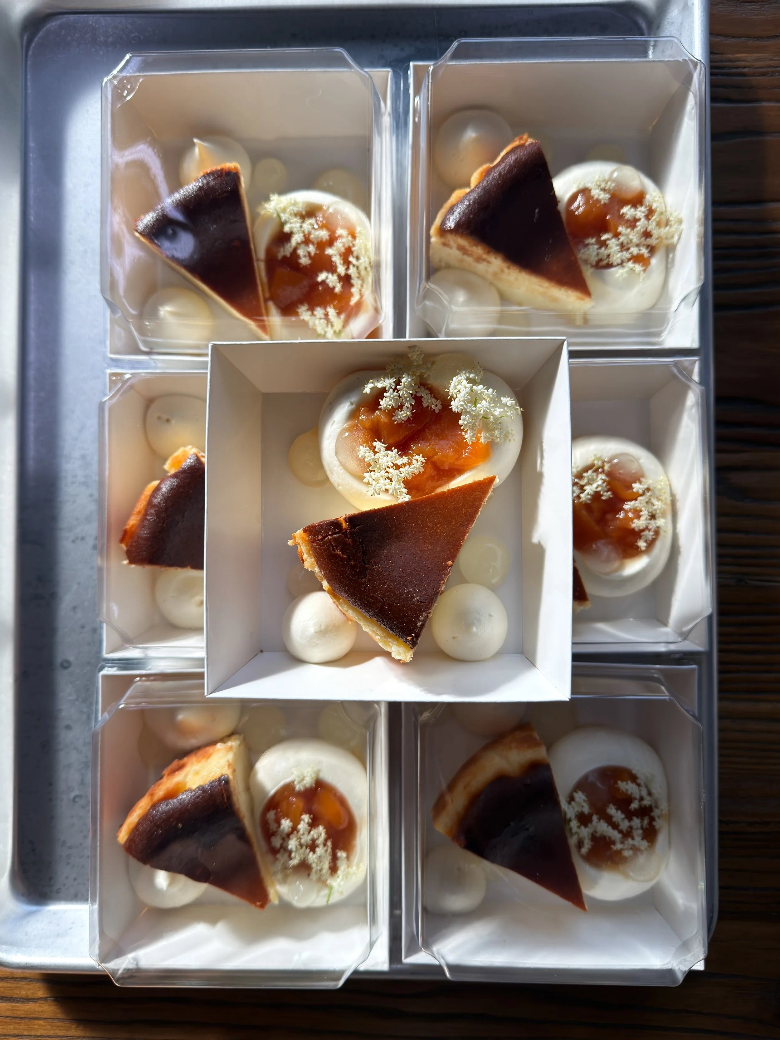 Close-up of individual dessert boxes containing a slice of basque cheesecake tart, topped with peach caramel sauce and elderflower garnish, arranged in a white box on a metallic tray from Florosa Bakery & Flower Farm, for purchase at JBird Supply.
