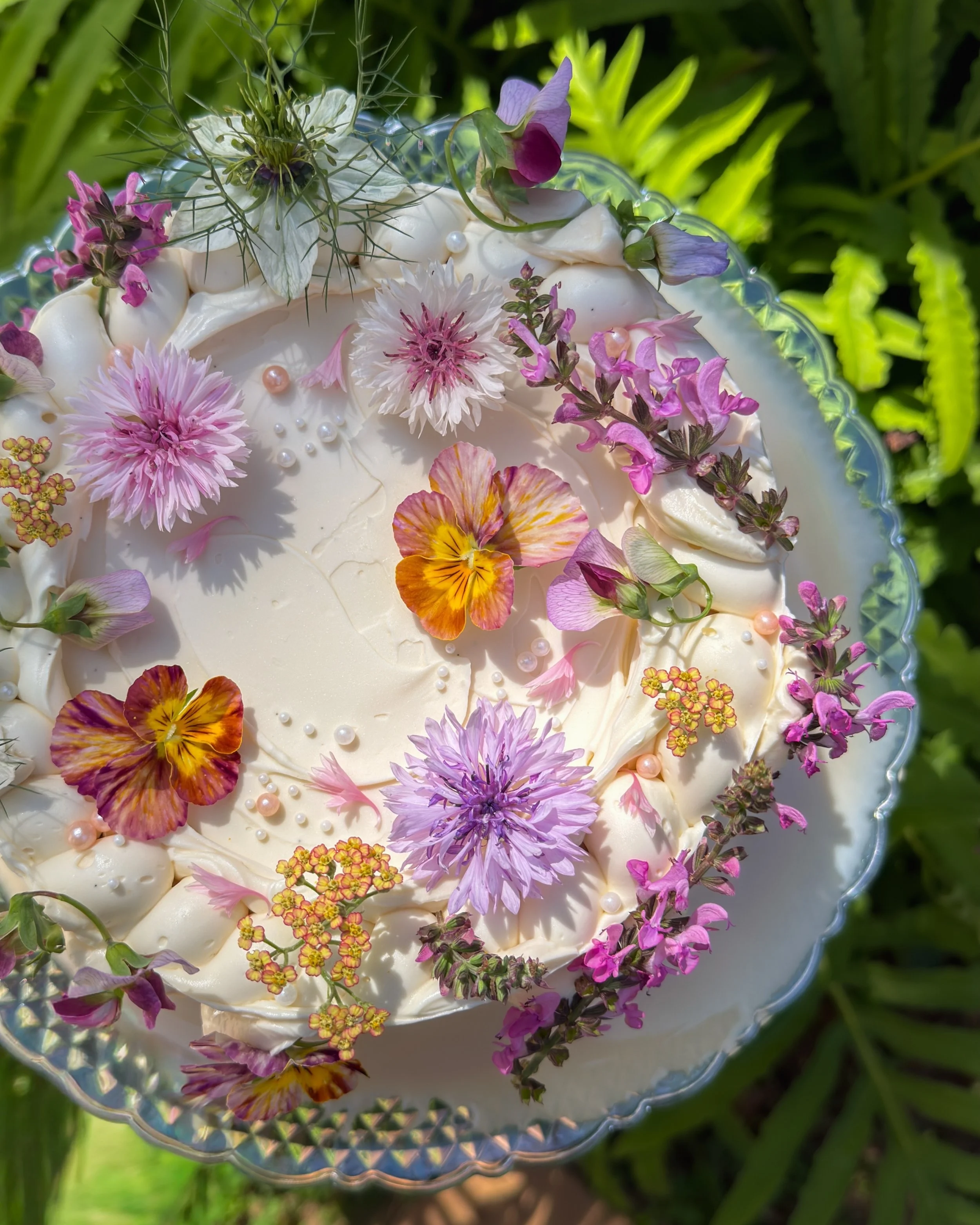 A tres leches cake from Florosa decorated with various colorful flowers and small pearl-like decorations on top, set outdoors on a tray in the garden.