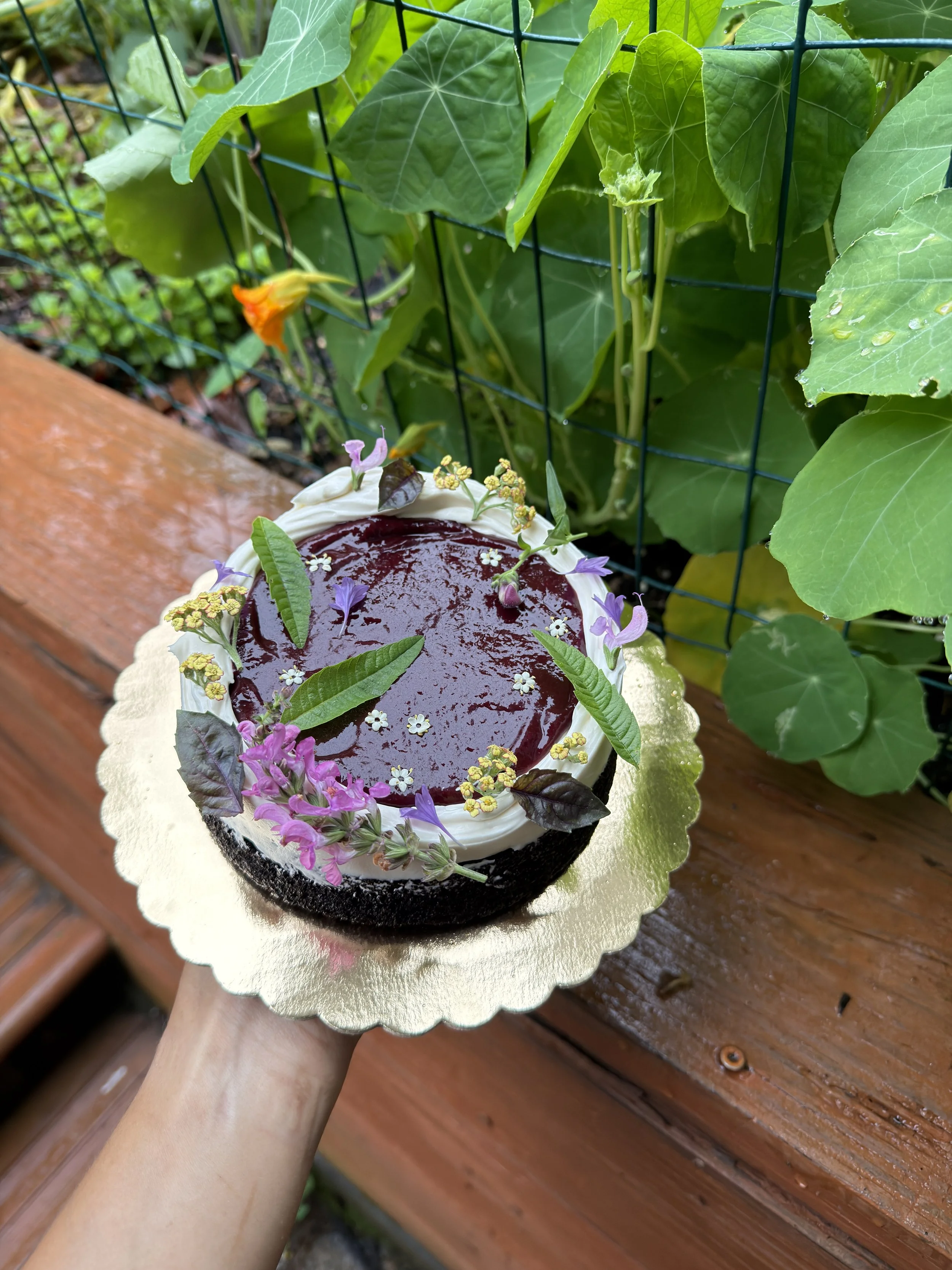 A person holding a vegan chocolate cake decorated with purple and white flowers, leaves, and berry-like fruit, with a glossy dark topping, outdoors on a wooden surface near leafy green plants from Florosa Bakery in Charlottesville, Virginia.