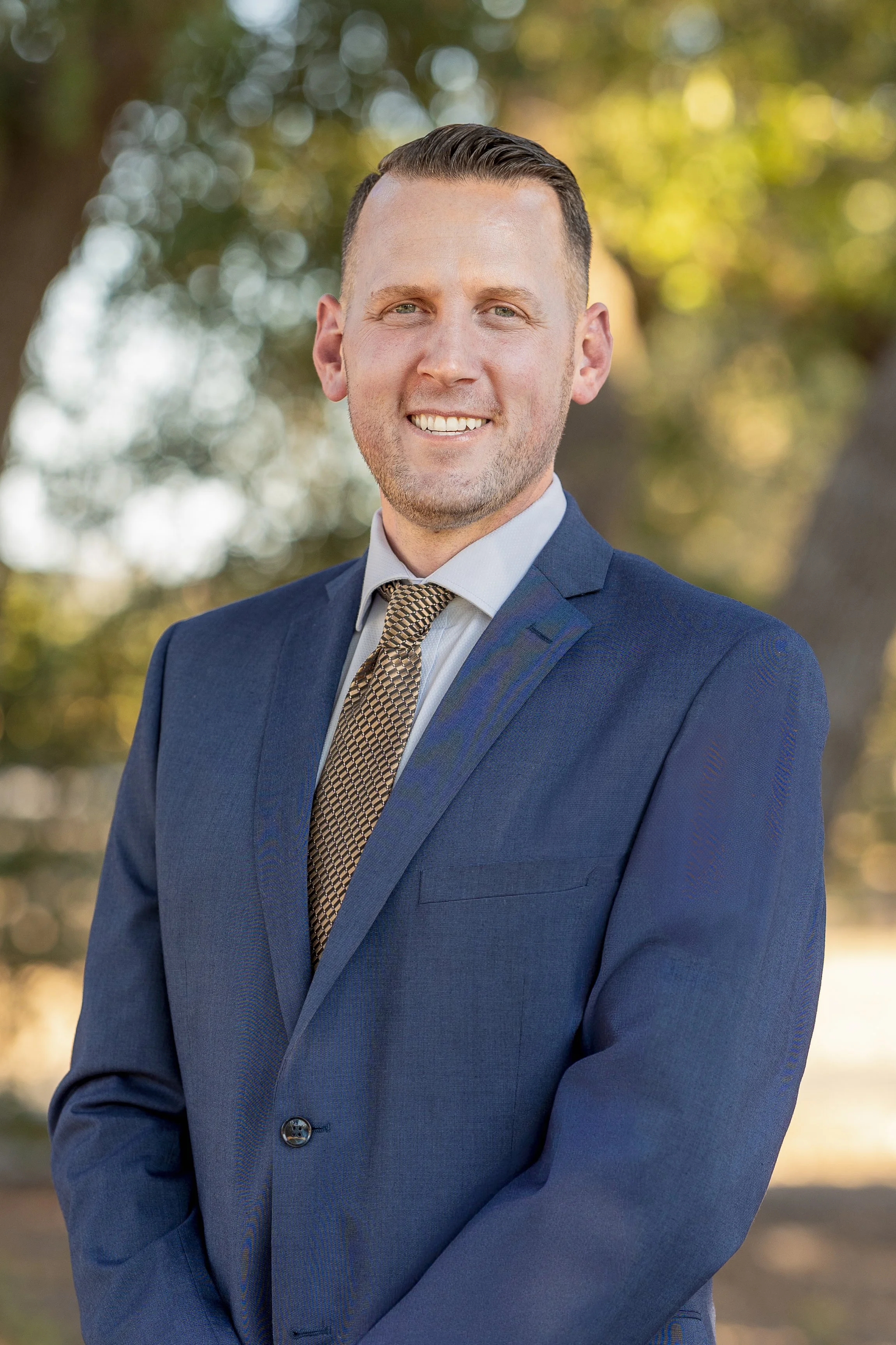 Portrait of a trustworthy man wearing a blue suit and patterned tie, smiling, with a blurred outdoor background of green trees and sunlight.