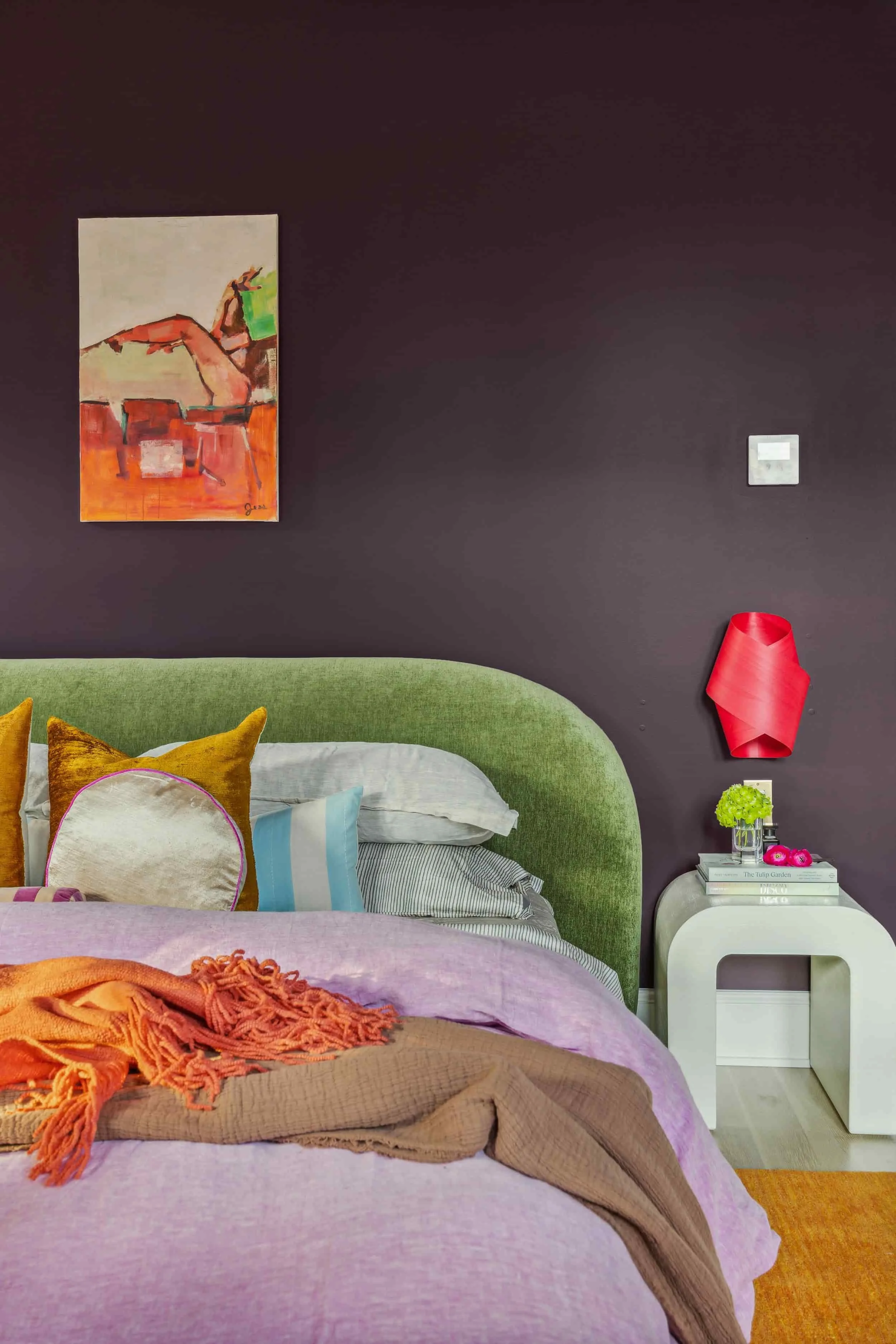 Colorful primary bedroom by Hannah Goetz with a curved green velvet headboard, lilac linen bedding, orange throw, plum walls and a white waterfall nightstand.